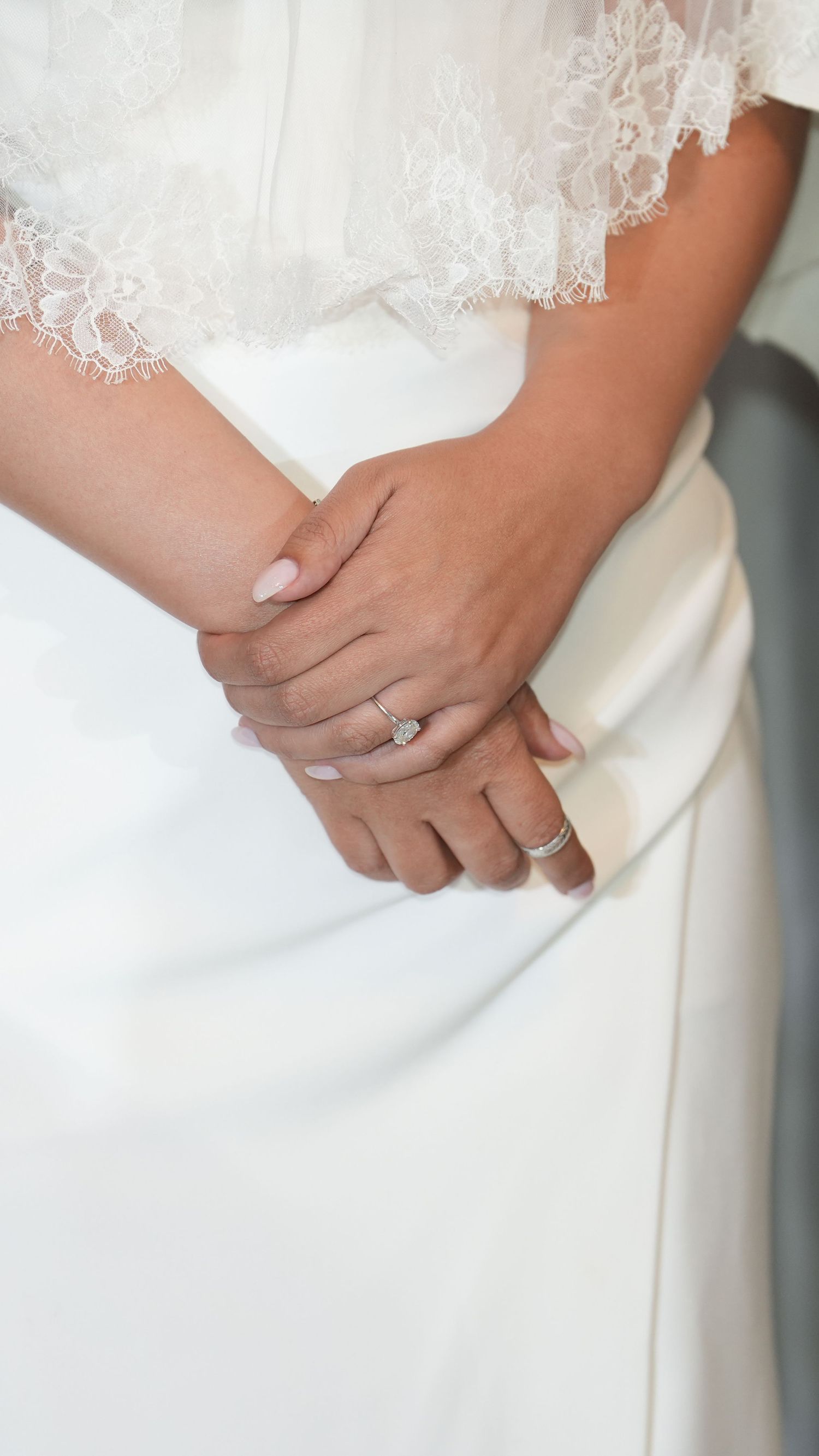 Close-up of a bride in a white lace wedding dress, clasping her hands showing a diamond engagement ring.