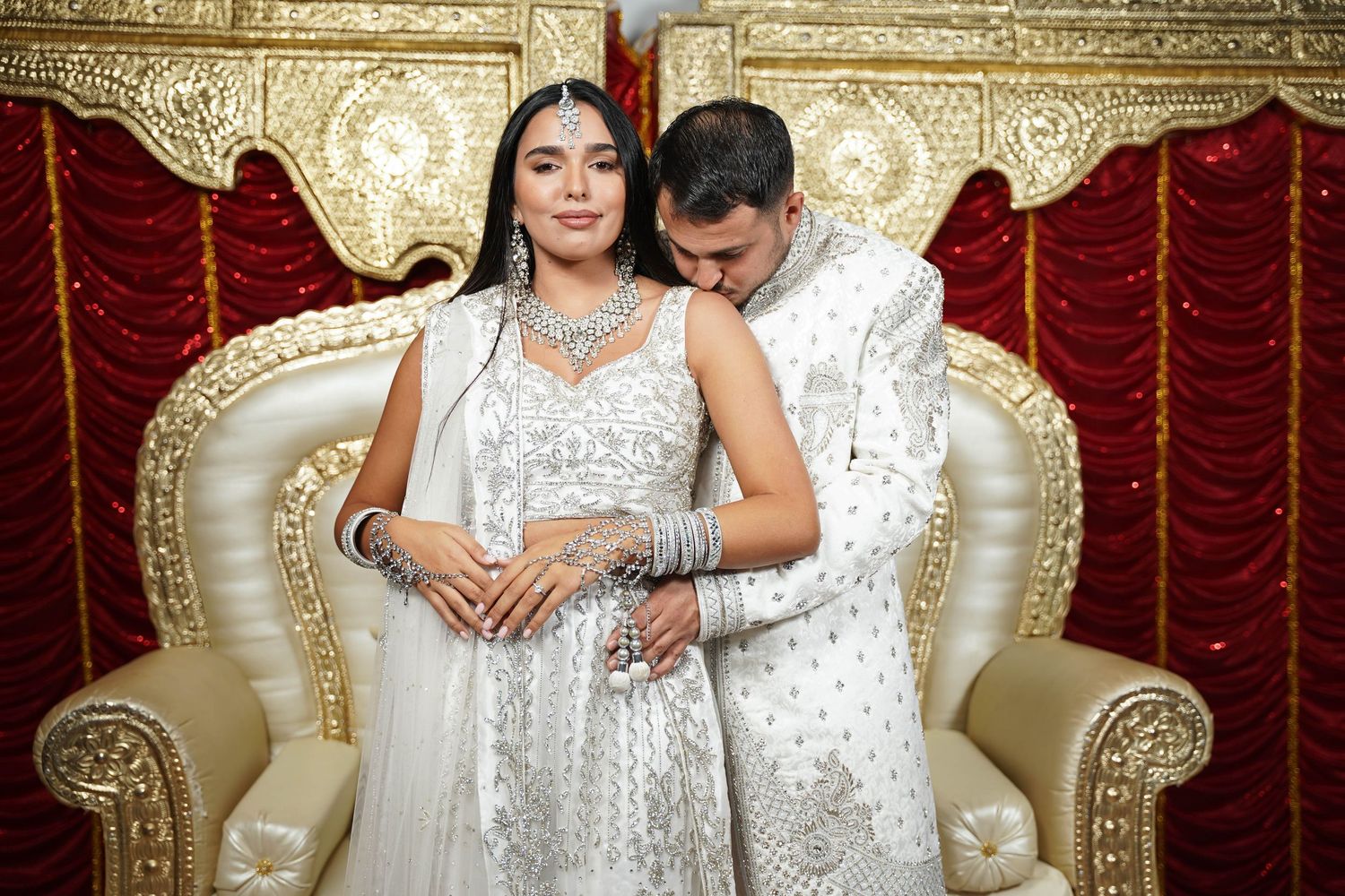 Elegant wedding couple in white attire posing on ornate gold throne.