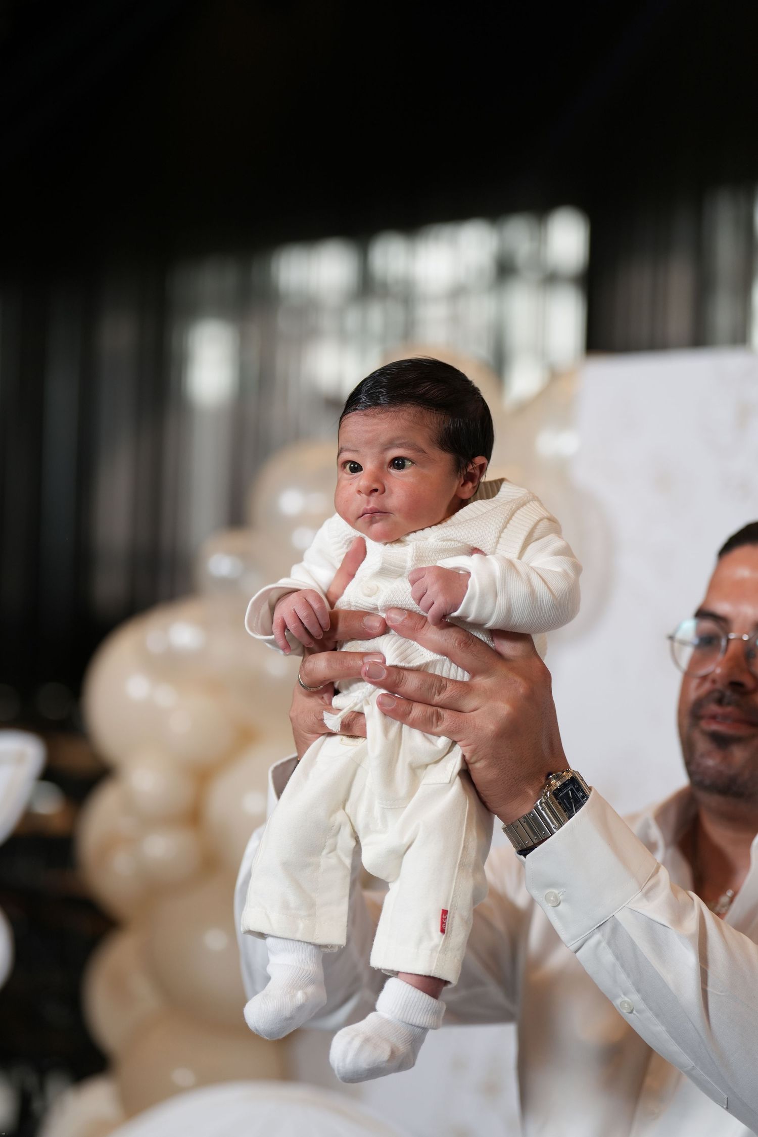 A newborn baby in a white outfit being held up by an adult man at a celebration with gold balloons.