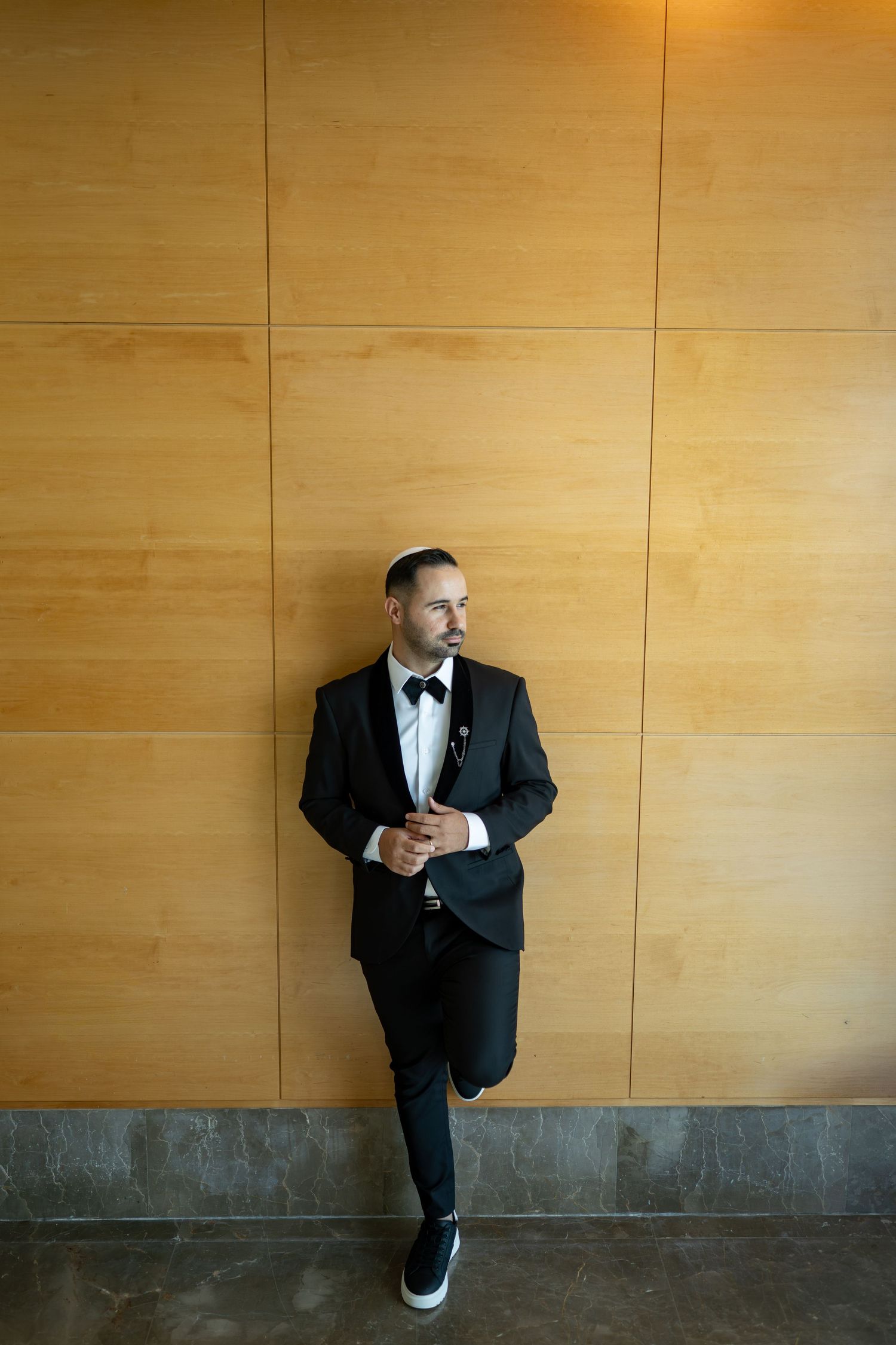 Stylish groom in black tuxedo and sneakers poses against wooden paneled wall.