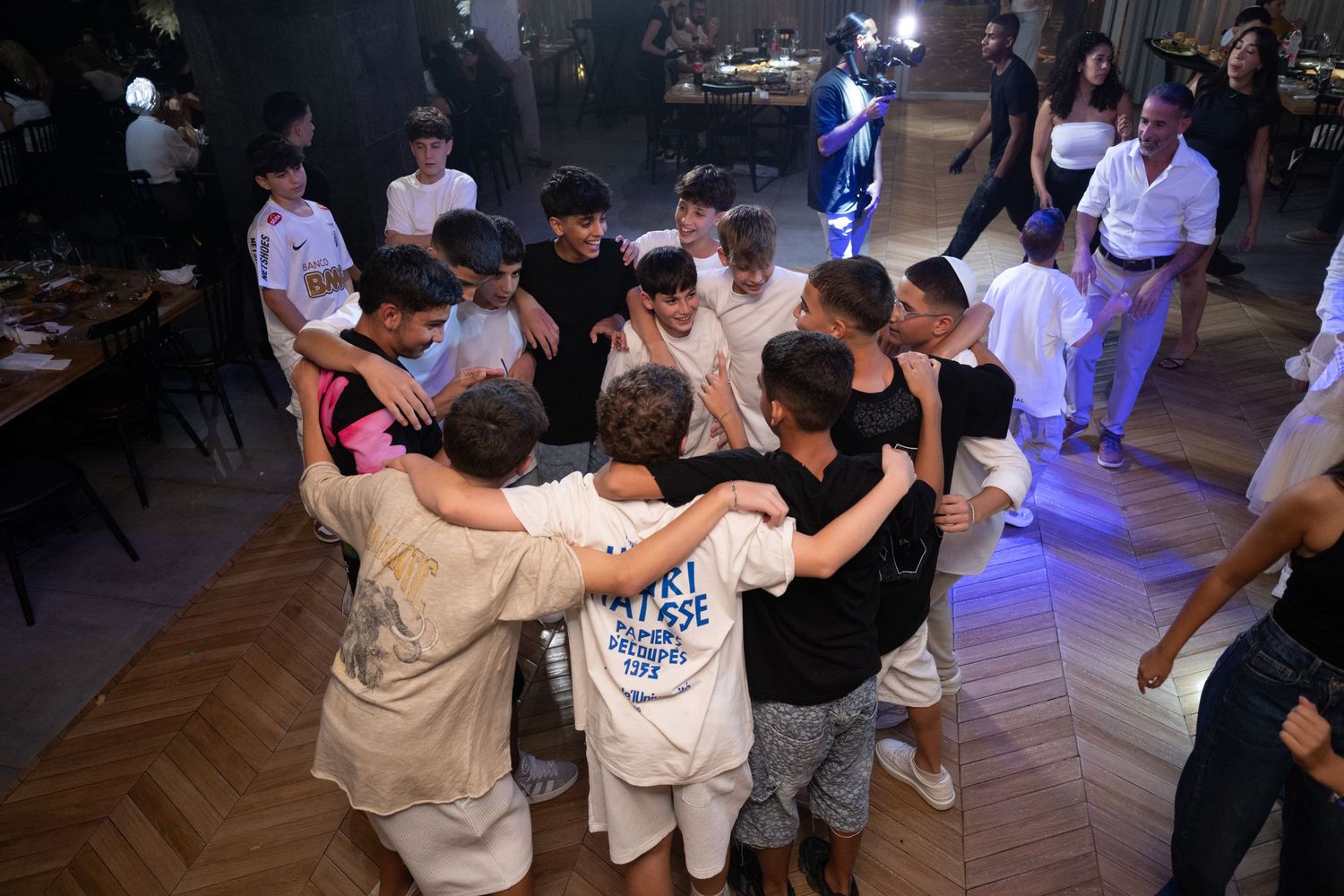 Large group dancing together in circle at indoor celebration with blue lighting.