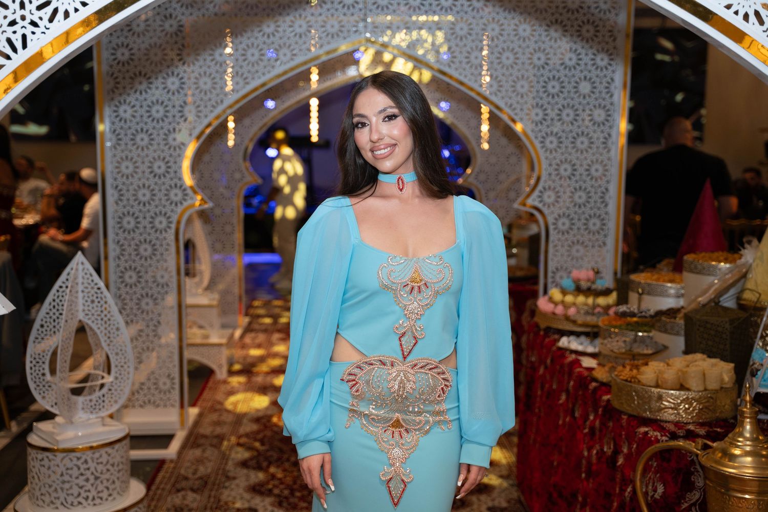 Person in turquoise outfit with gold embellishments poses at elegant indoor event with ornate decor and lighting.