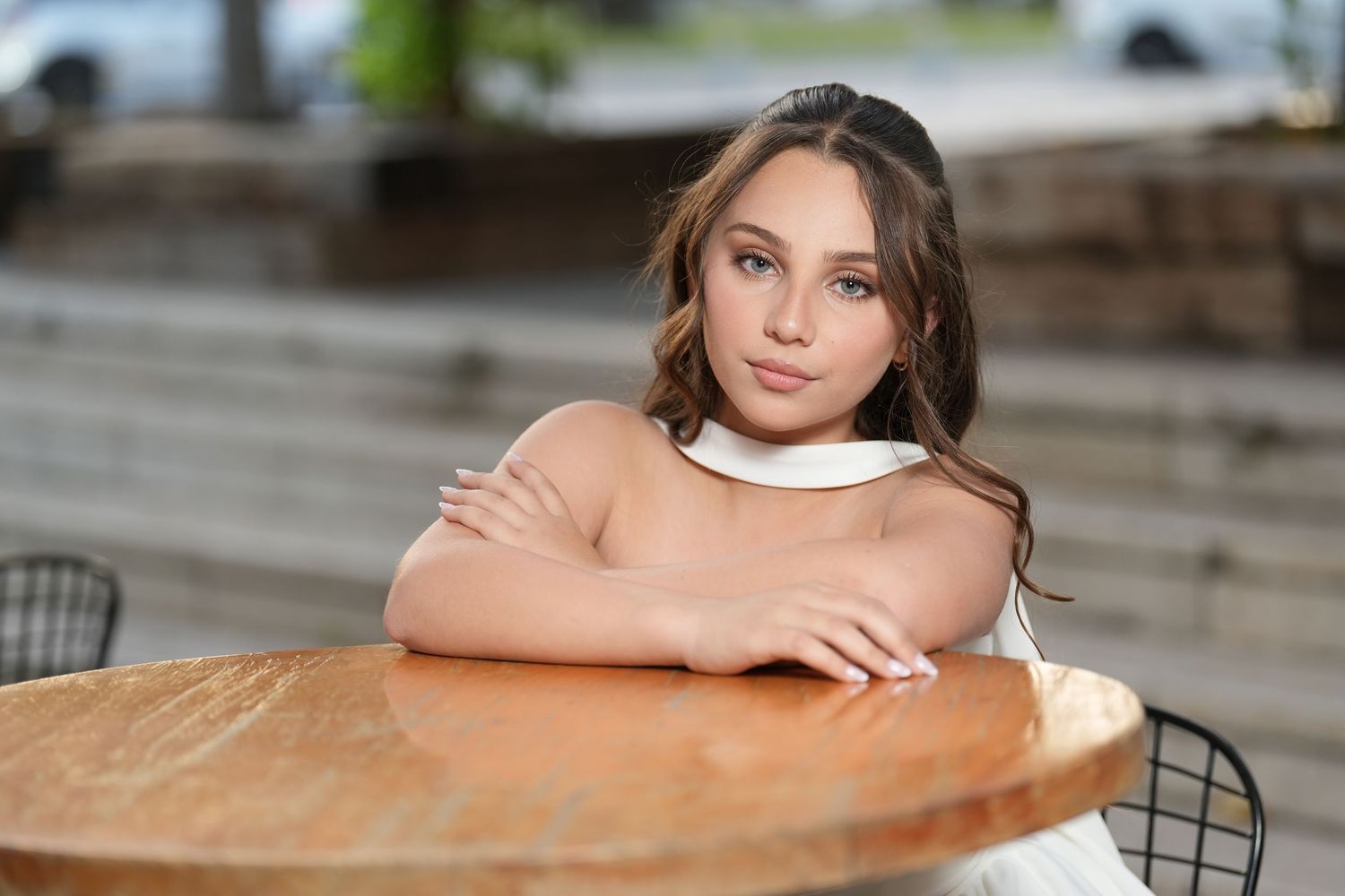Young woman with wavy hair leans on a wooden table outdoors, wearing a white top, gazing thoughtfully at the camera.