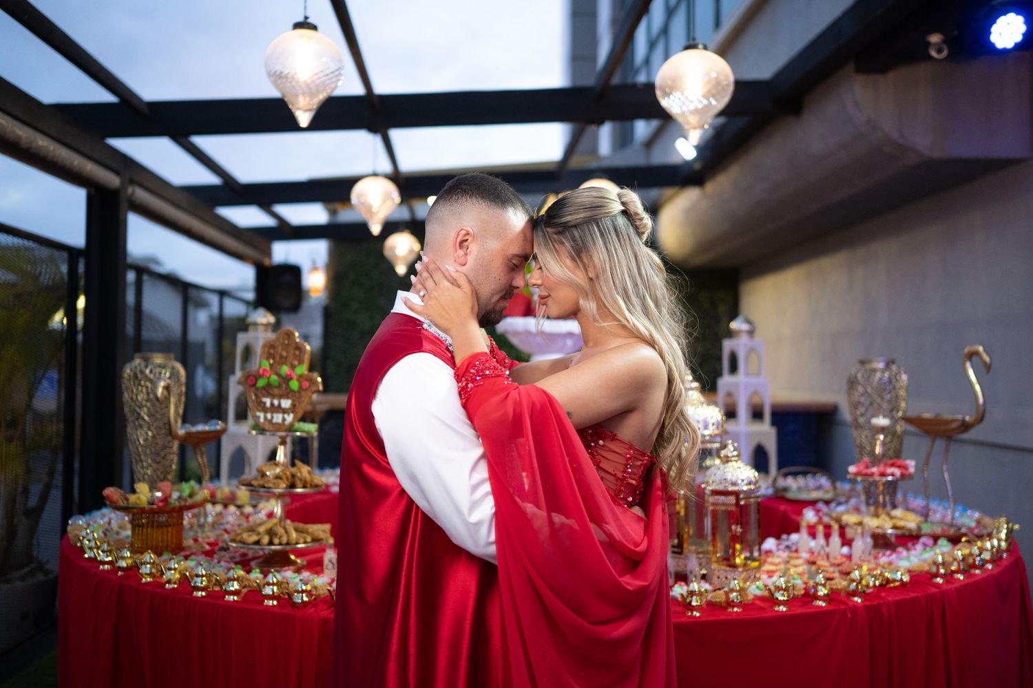 Couple in matching red outfits embracing near a decorated dessert table with golden accents and elegant lighting.