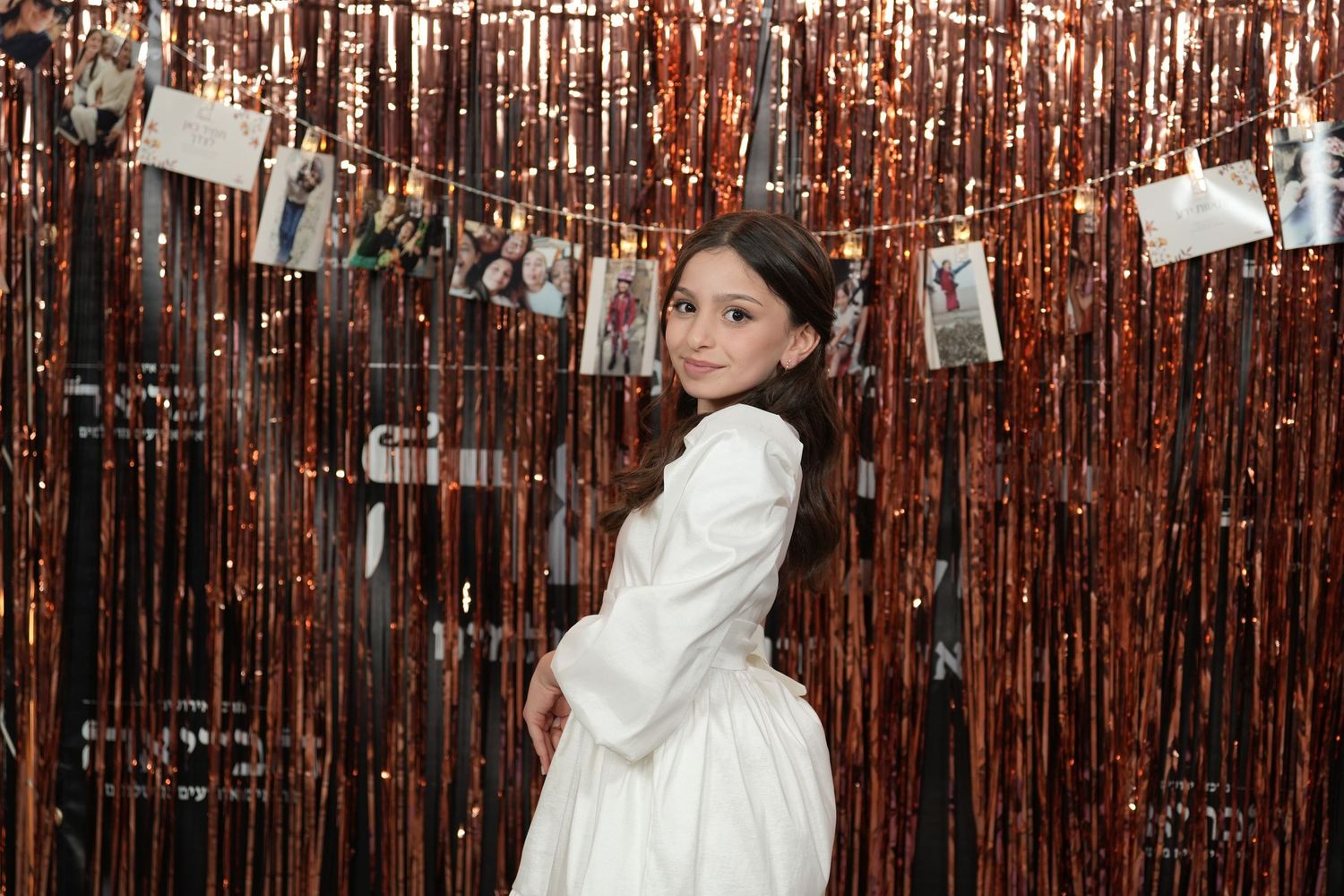 A child in a white dress poses in front of a rose gold tinsel backdrop with photos hanging on string.