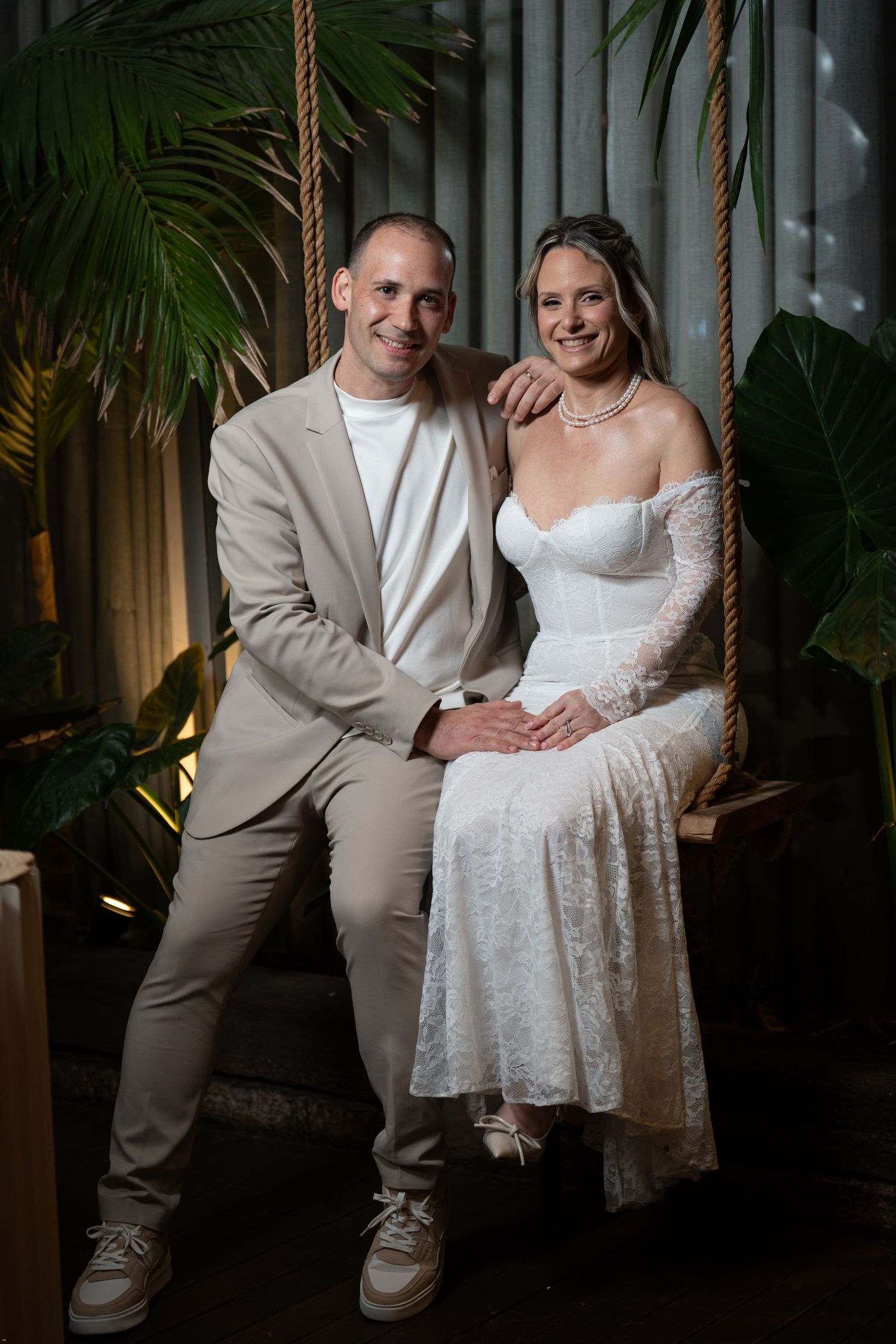 A couple in wedding attire sits together on a decorative swing in an elegant indoor setting with tropical plants.