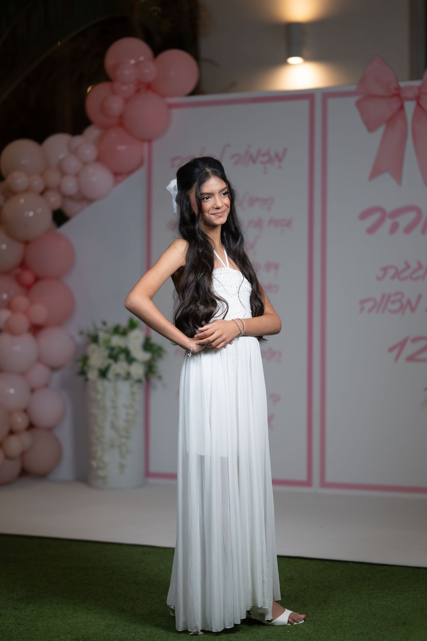 An elegant white maxi dress displayed in a room with pink balloon decorations and floral accents.
