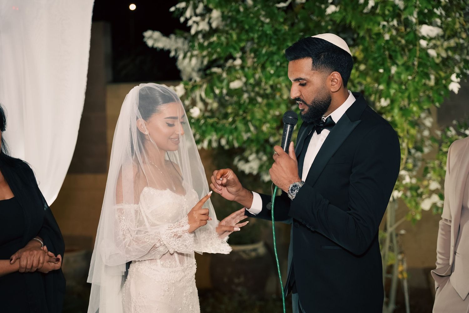 Bride and groom share a romantic moment during their wedding ceremony under hanging lights and greenery.
