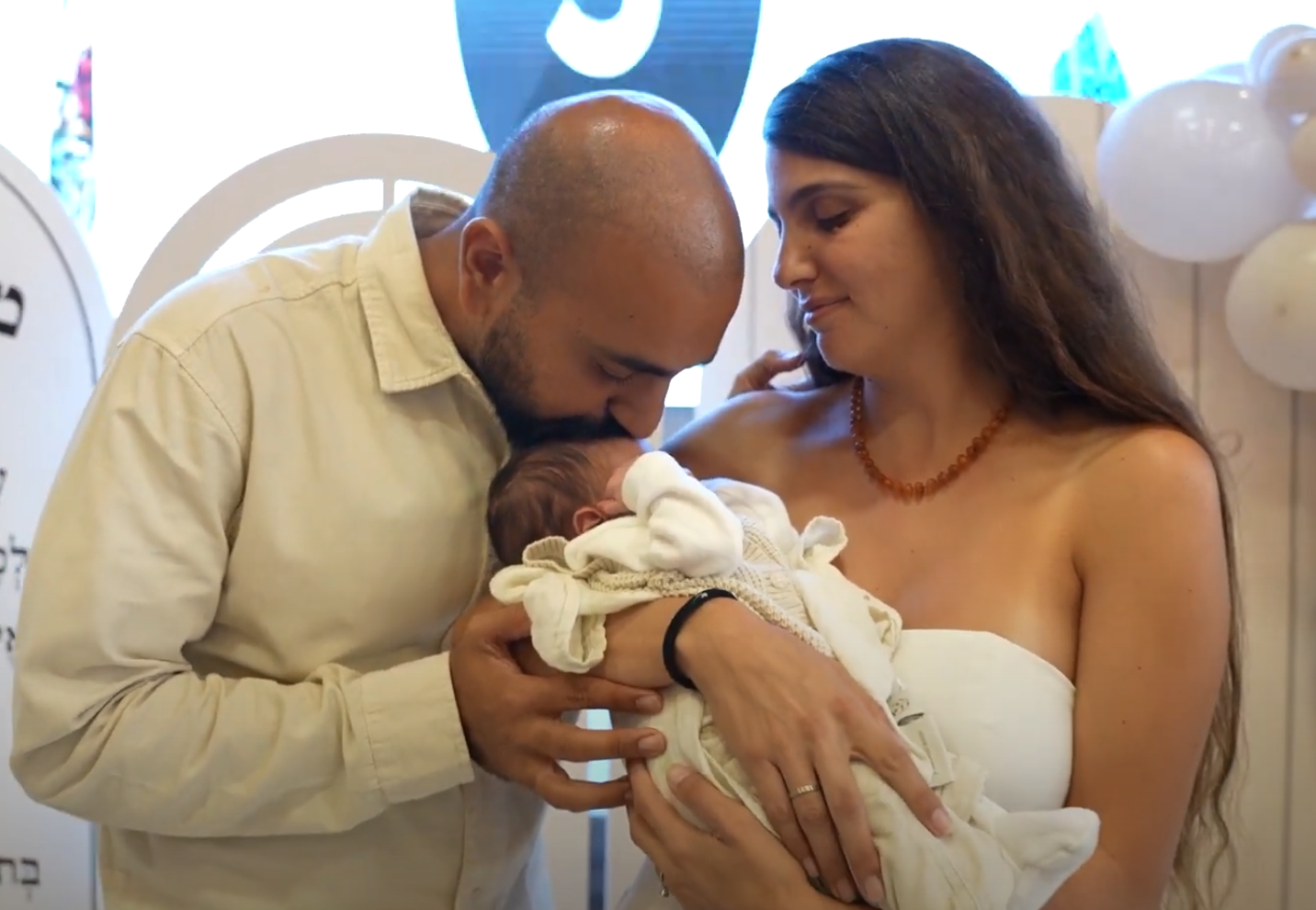 A tender moment as new parents gaze at their swaddled newborn baby under blue and white balloons.
