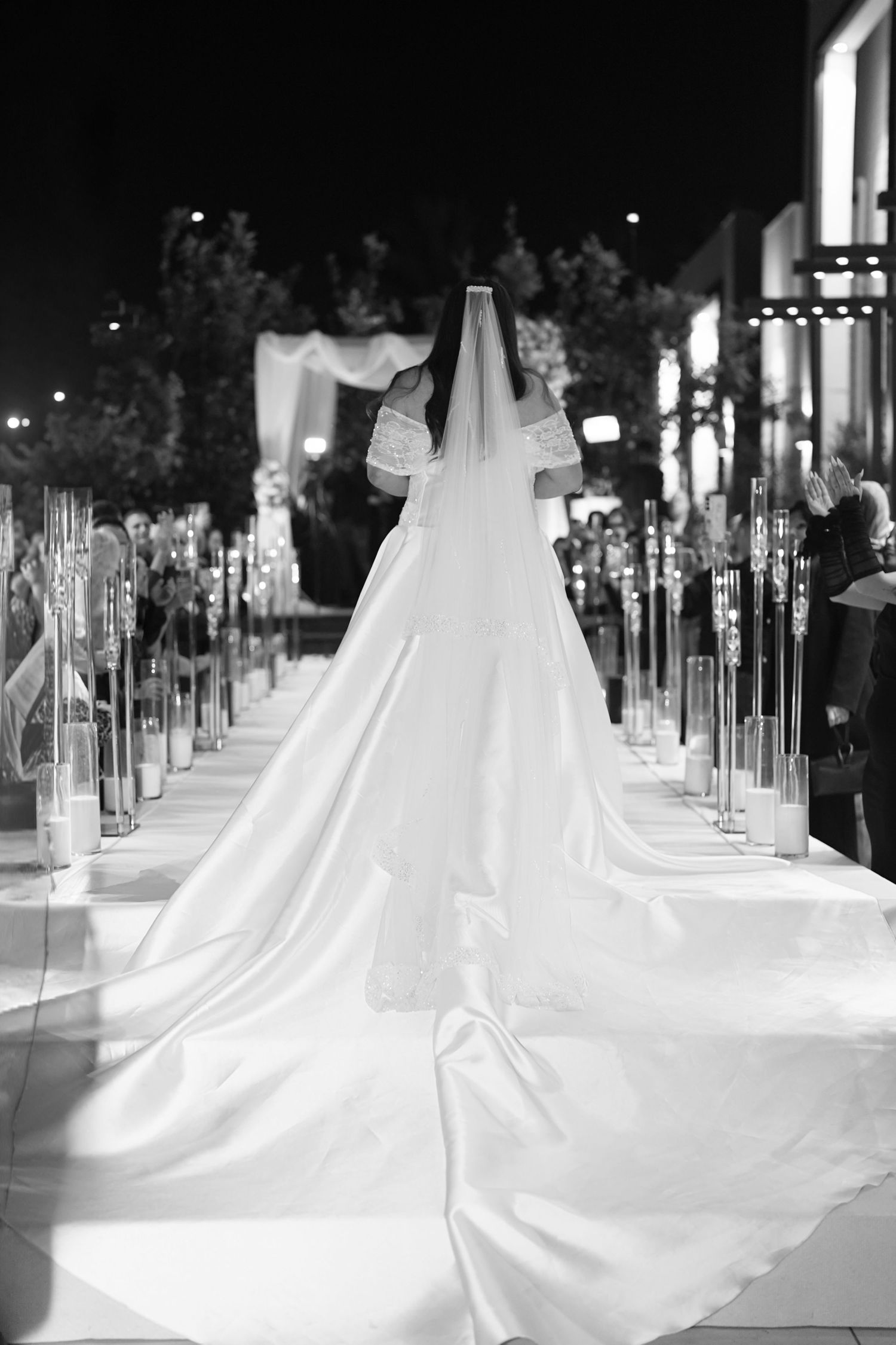 Black and white photo of a bride walking down the aisle in a stunning gown with a long train and veil at night.