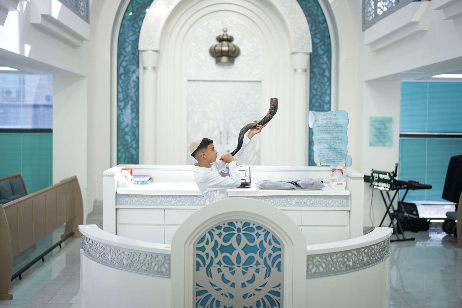 Man blowing a shofar at a decorative white reception desk in an elegant teal and white synagogue interior.
