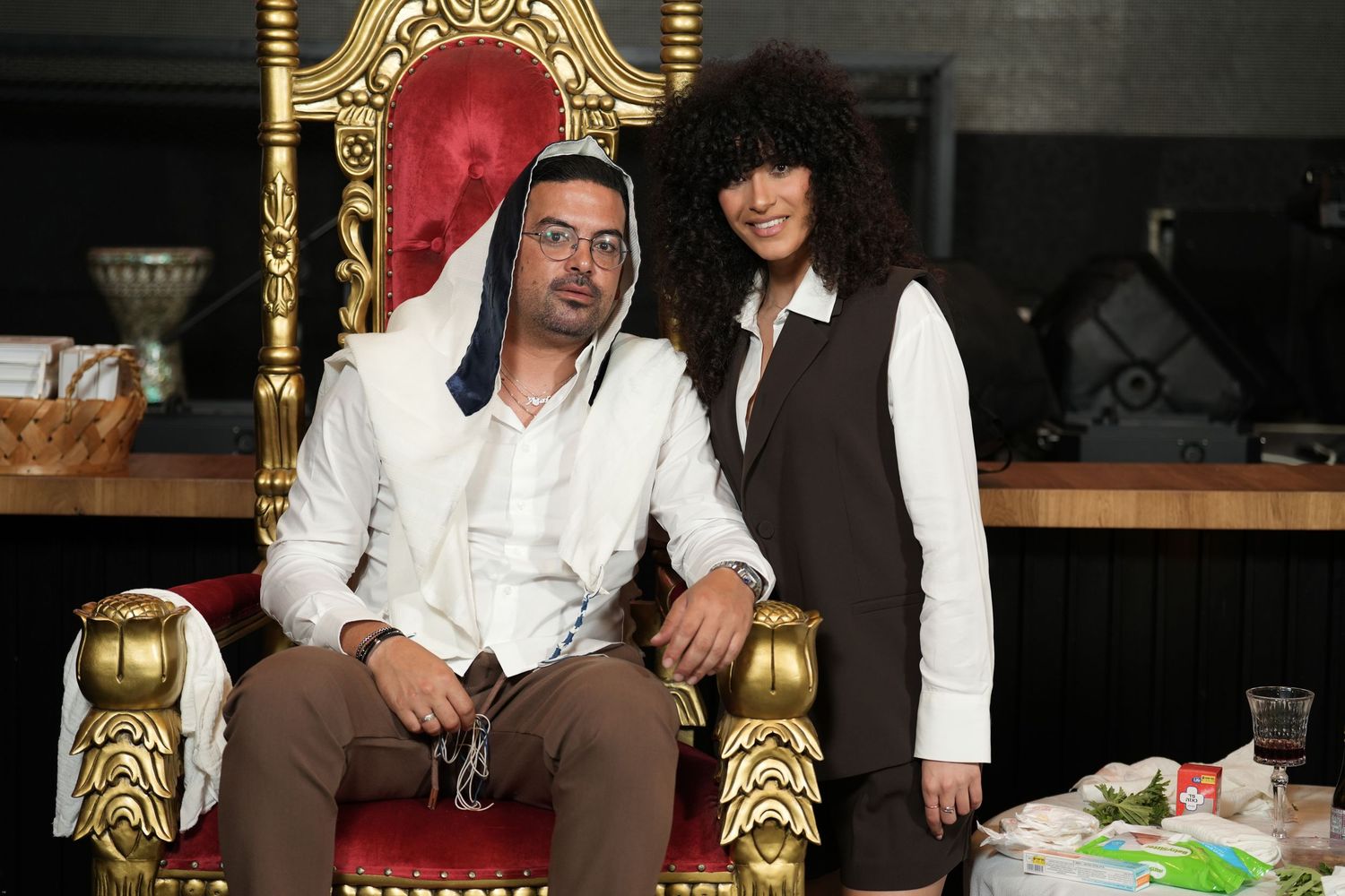 Two people posing on a ornate gold and red throne chair in a dimly lit room.