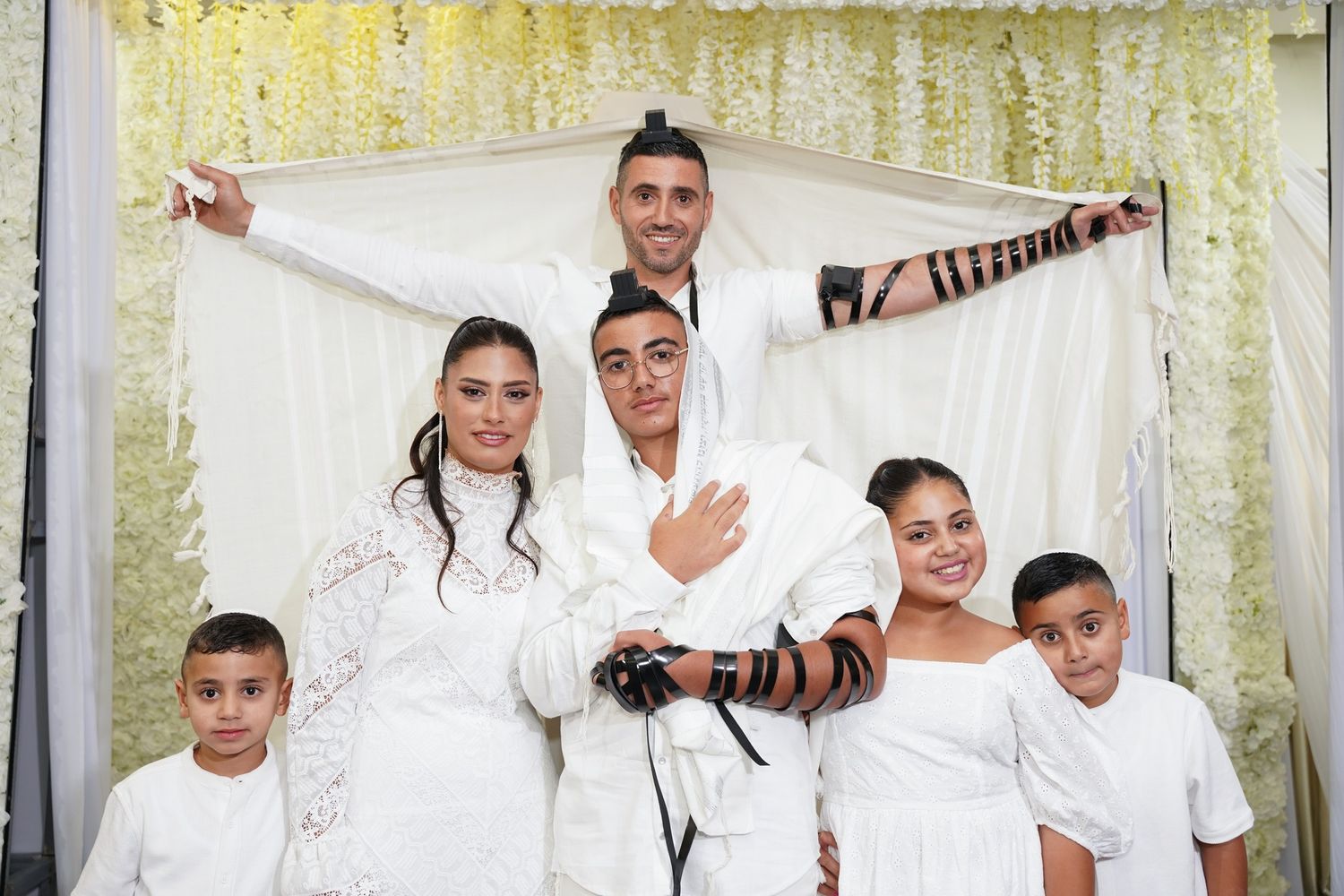 Family group photo with traditional religious attire and prayer shawl against floral backdrop.