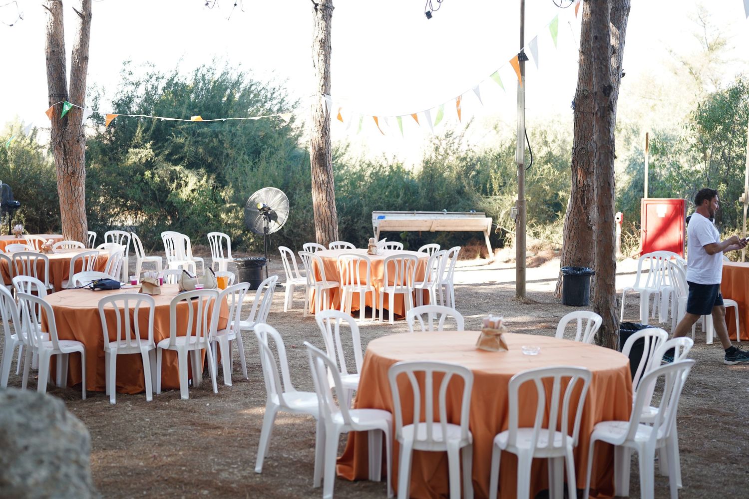 Outdoor party setup with white plastic chairs and orange tablecloths arranged under trees.