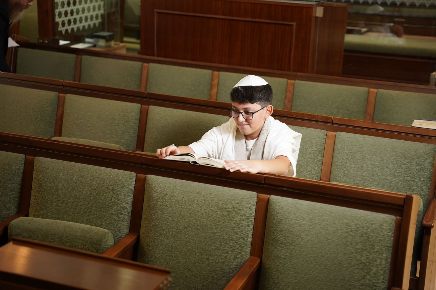 Someone wearing a white shirt and cap sits alone in an auditorium with green upholstered seats.
