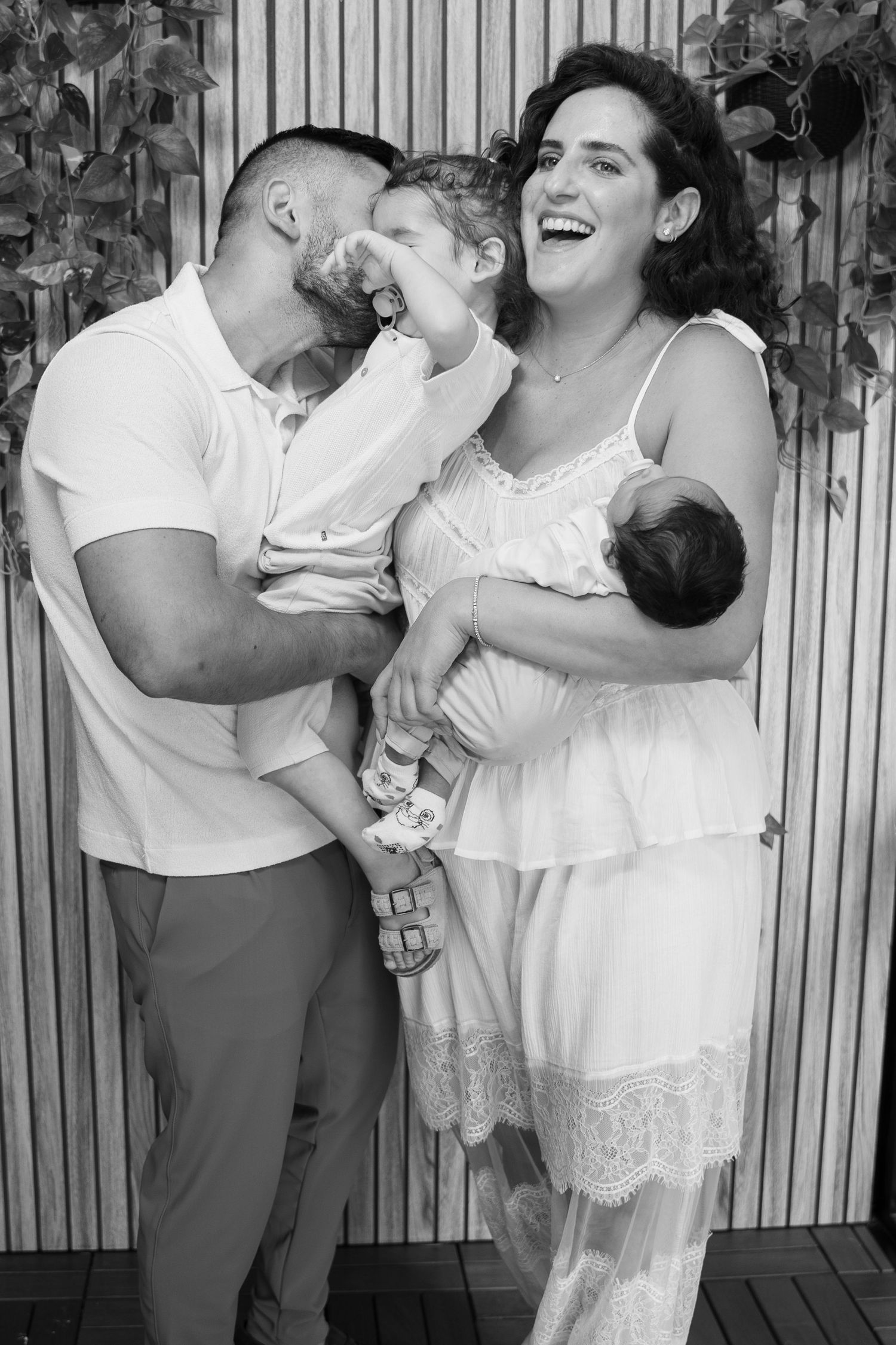 Black and white family photo of parents laughing and kissing toddler while mother holds newborn baby outdoors.