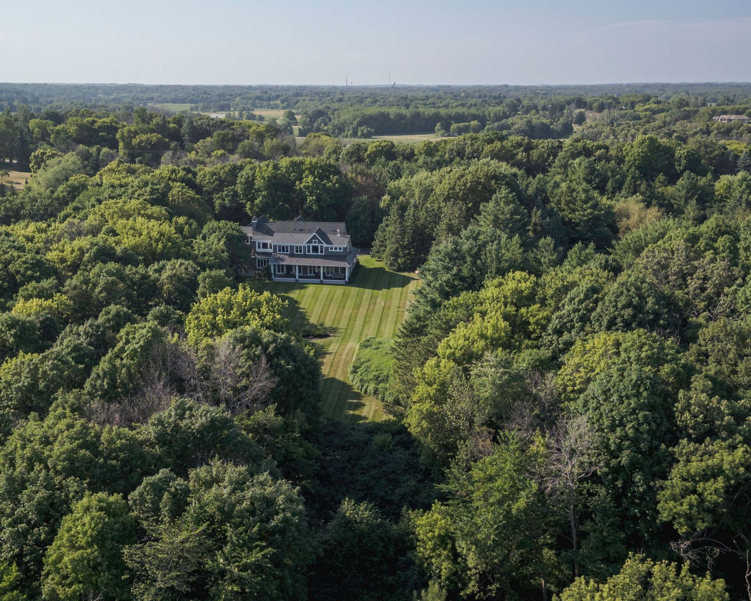 Aerial view of a large estate home surrounded by lush green forest and manicured lawn in a rural setting.