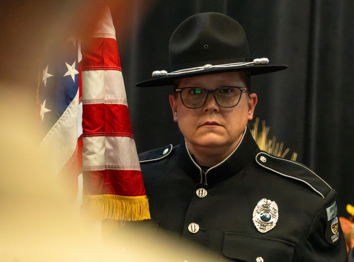 Law enforcement officer in formal uniform with hat stands next to American flag.