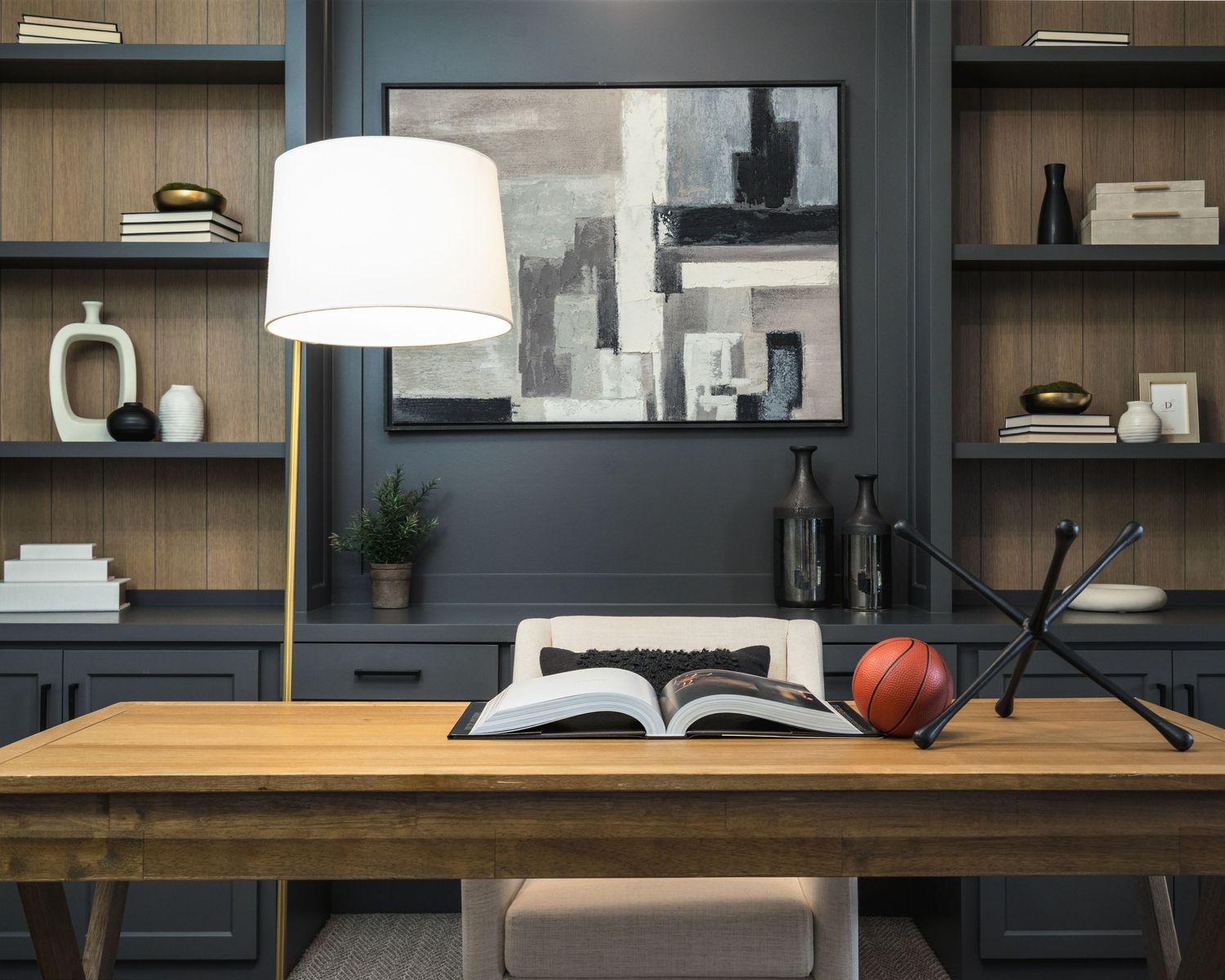 A modern home office features dark built-in cabinets, wooden shelving, and white decor accents against a gray wall.