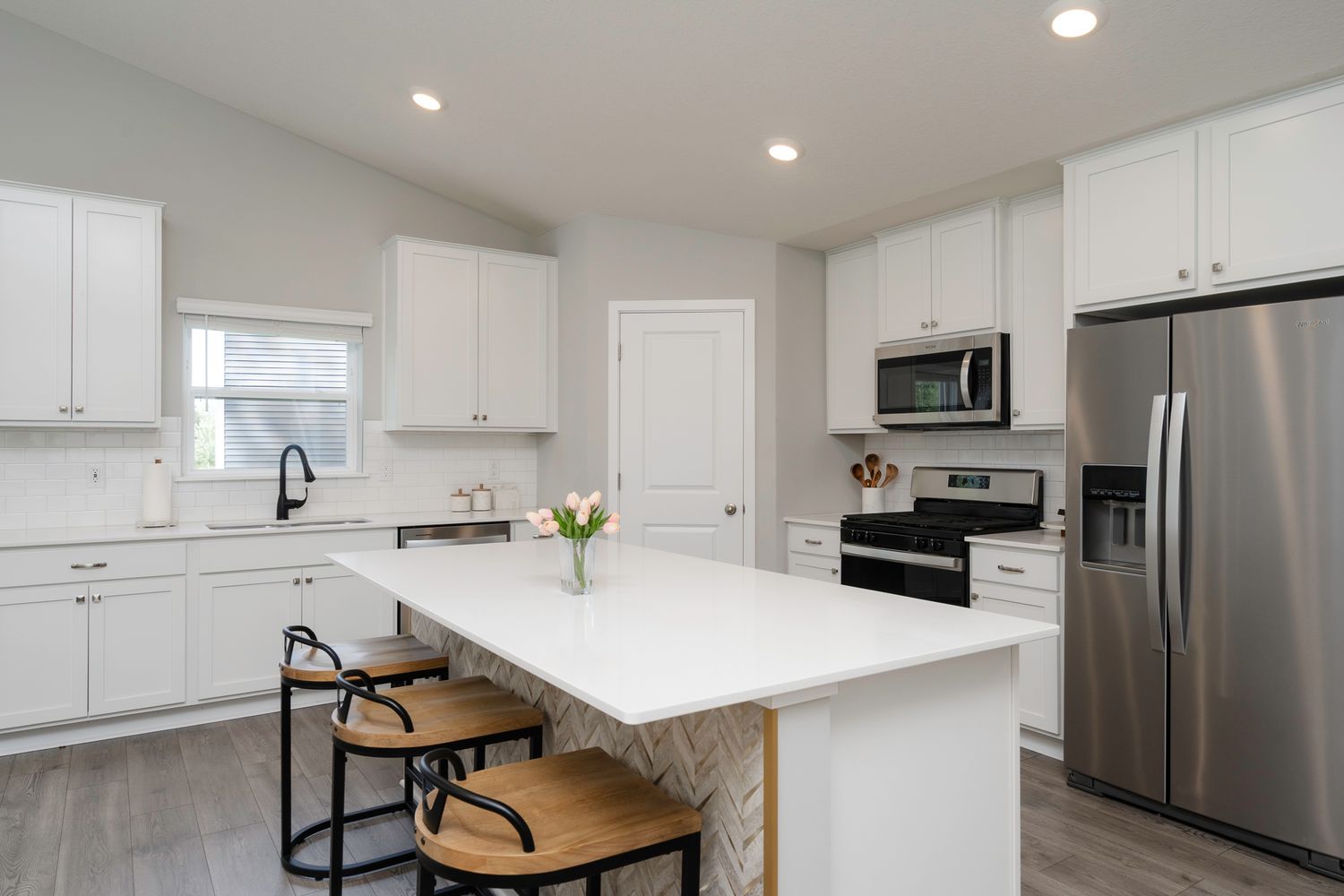 Modern white kitchen with island, quartz countertops, stainless steel appliances, and bar stools on light hardwood floors.