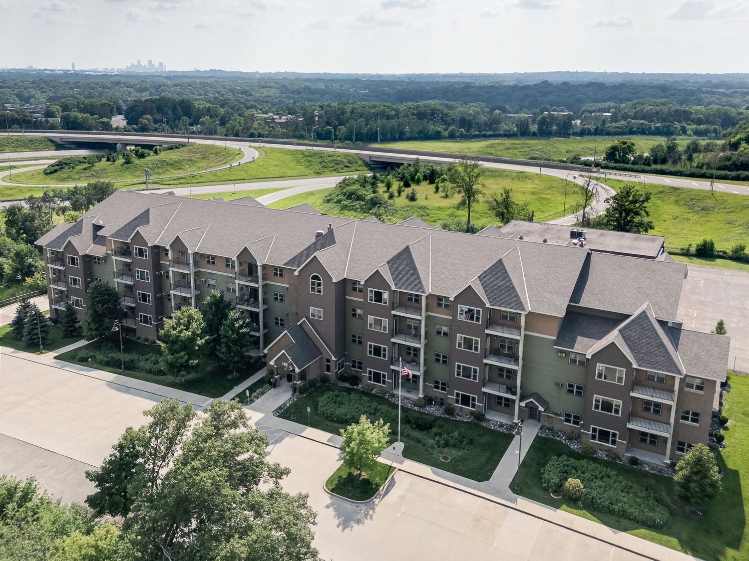Aerial view of a multi-story brick apartment complex surrounded by lush trees near a highway overpass.