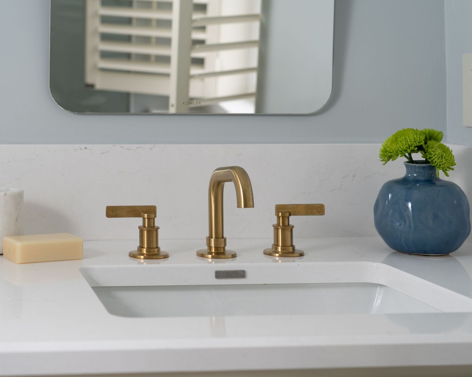 Modern bathroom vanity with gold brushed brass widespread faucet, white undermount sink, and blue vase with green flowers.