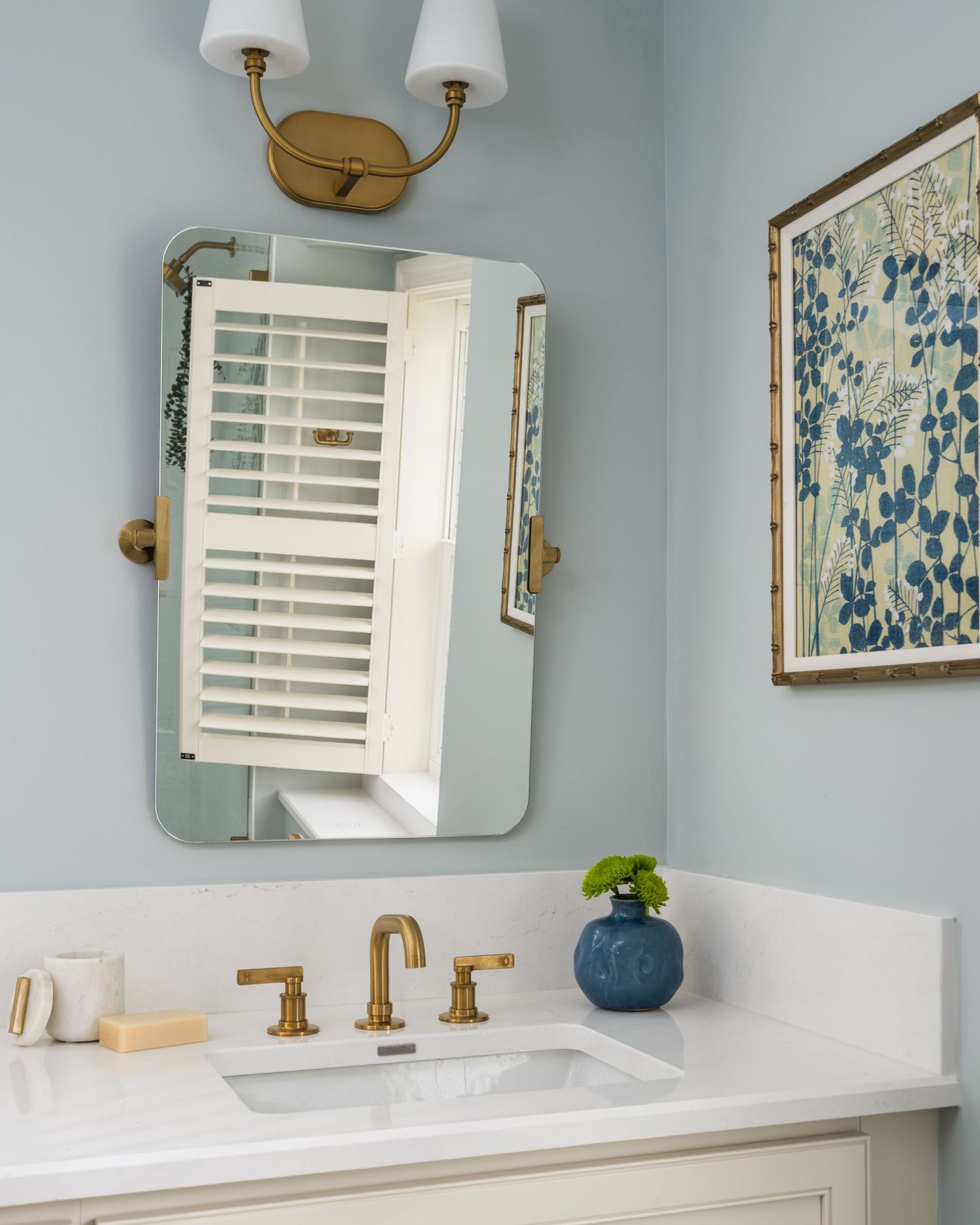 Light blue bathroom with gold faucet, white vanity, beveled mirror, brass sconce, and blue floral artwork on wall.