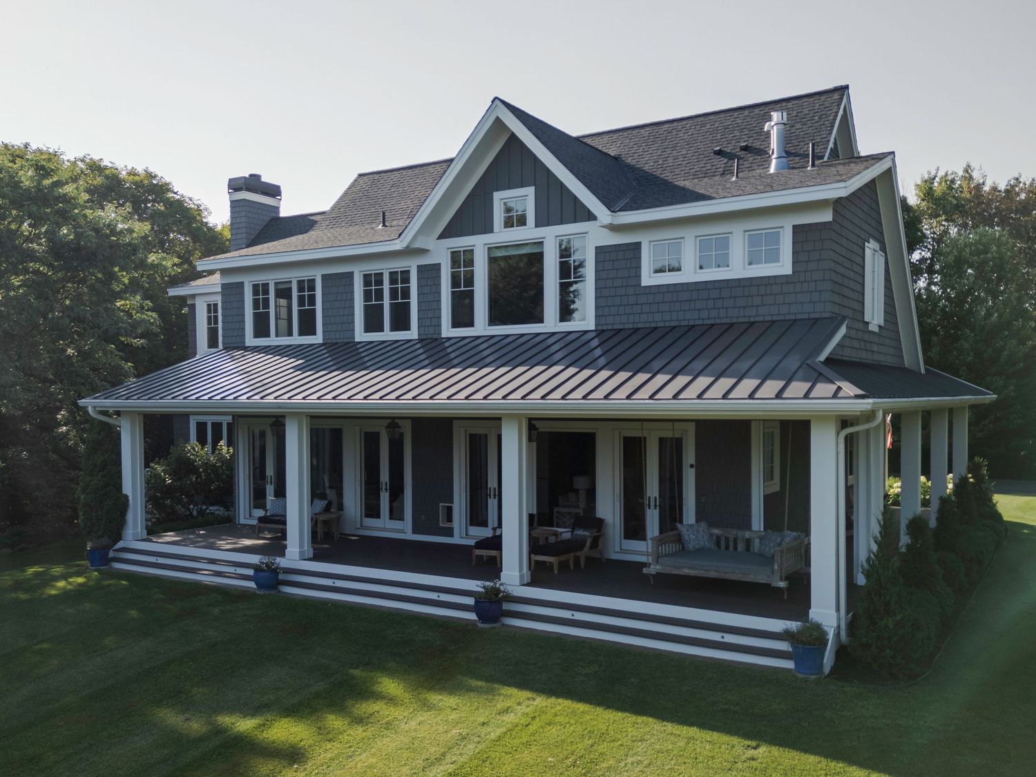 Large gray craftsman-style home with wrap-around porch, white columns, and metal roof surrounded by lush green lawn.