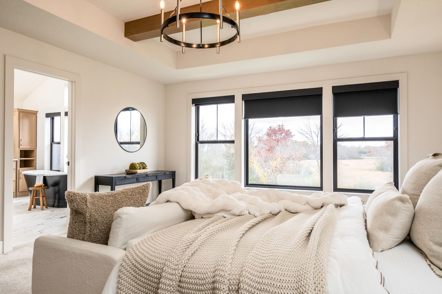Bright modern bedroom with tray ceiling, black-framed windows, neutral bedding, and a vanity area with round mirror.