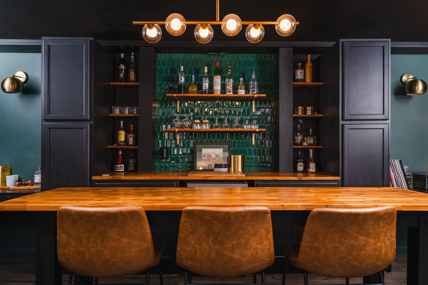 Modern home bar with teal walls, leather barstools, and illuminated glass shelving displaying liquor bottles.