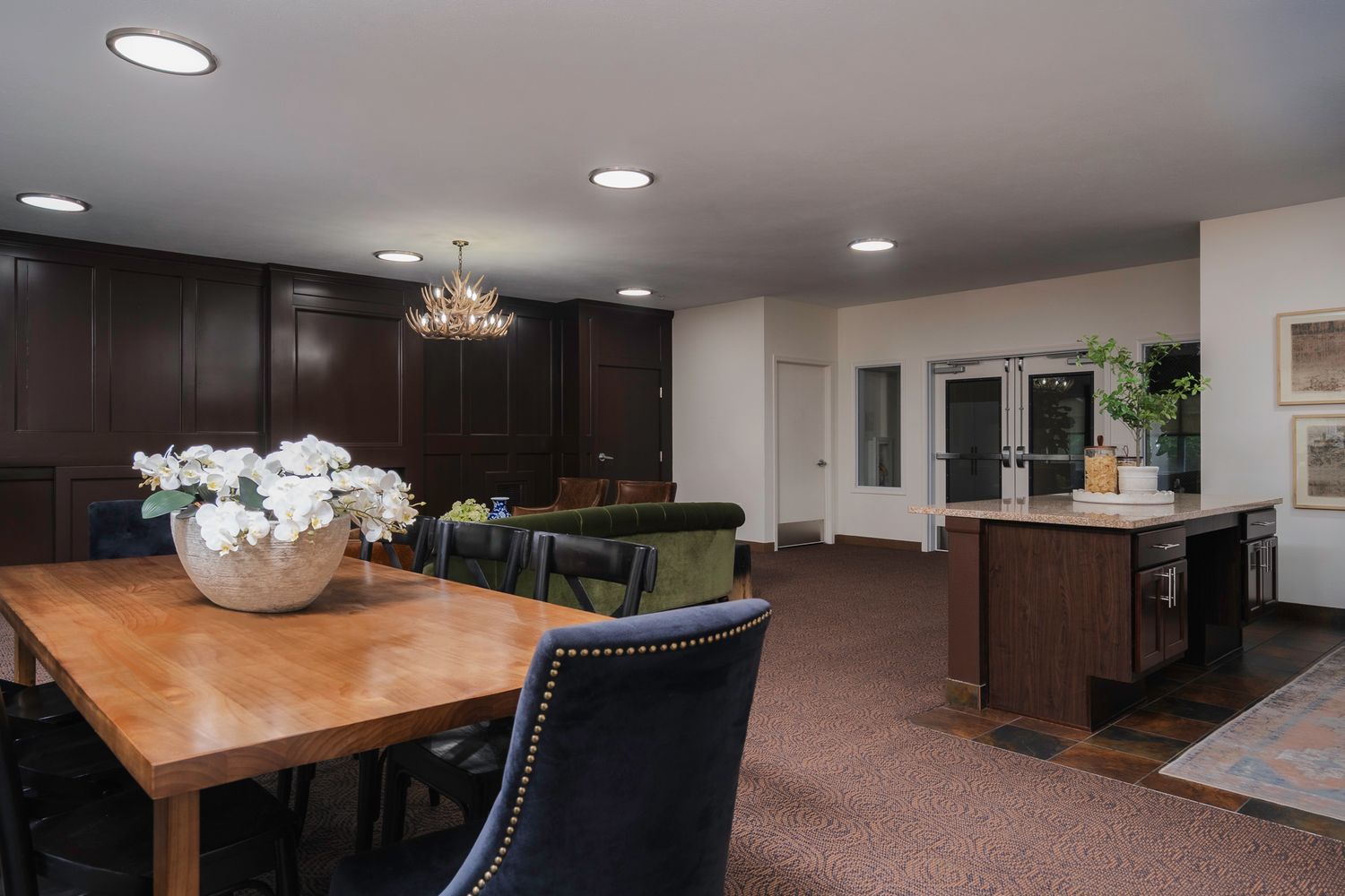 Elegant dining room with wood table, navy chairs, chandelier, dark cabinetry, and open kitchen area with carpet flooring.