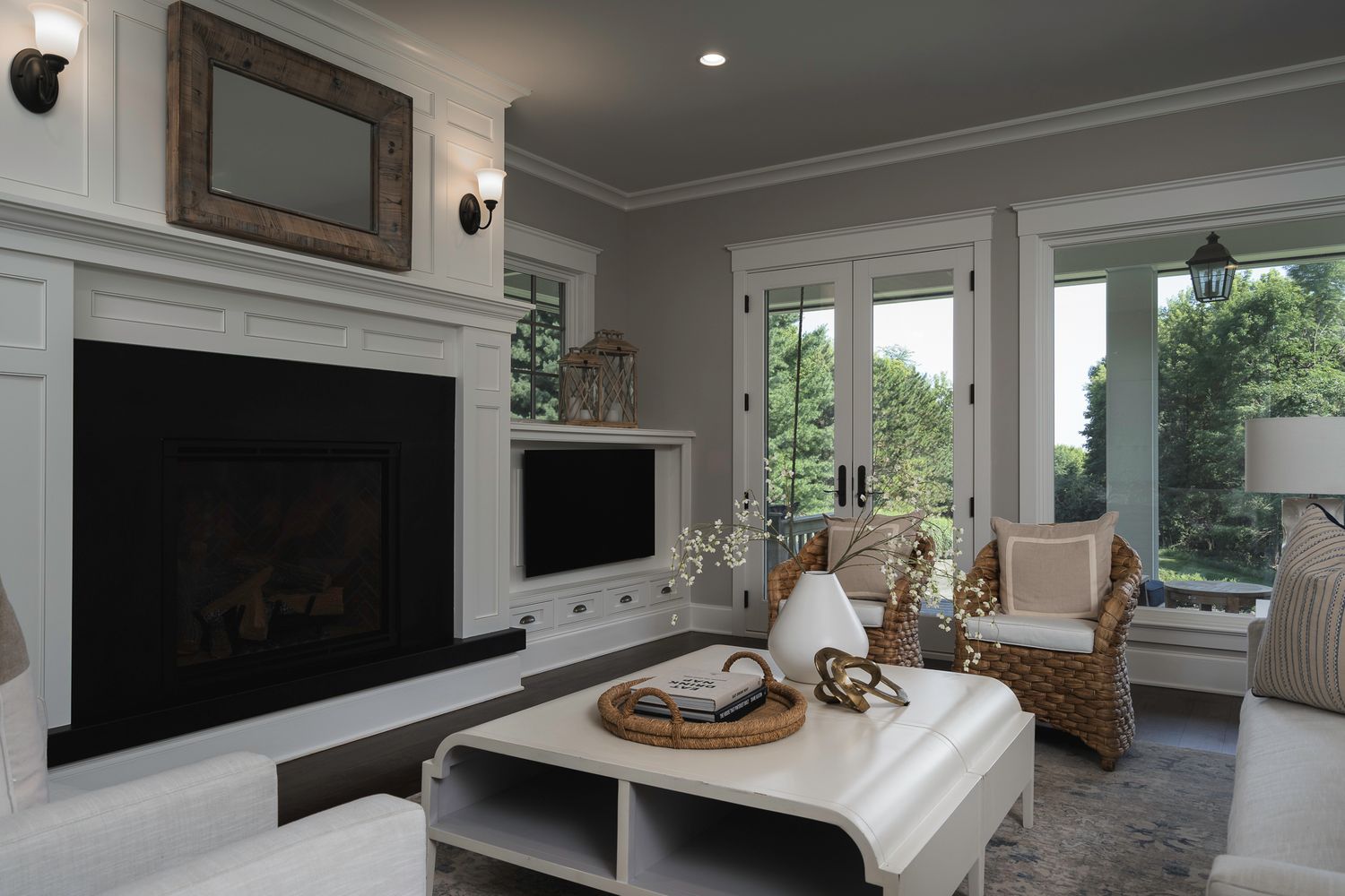 Elegant living room with white fireplace, built-ins, wicker chairs, and large windows overlooking greenery.