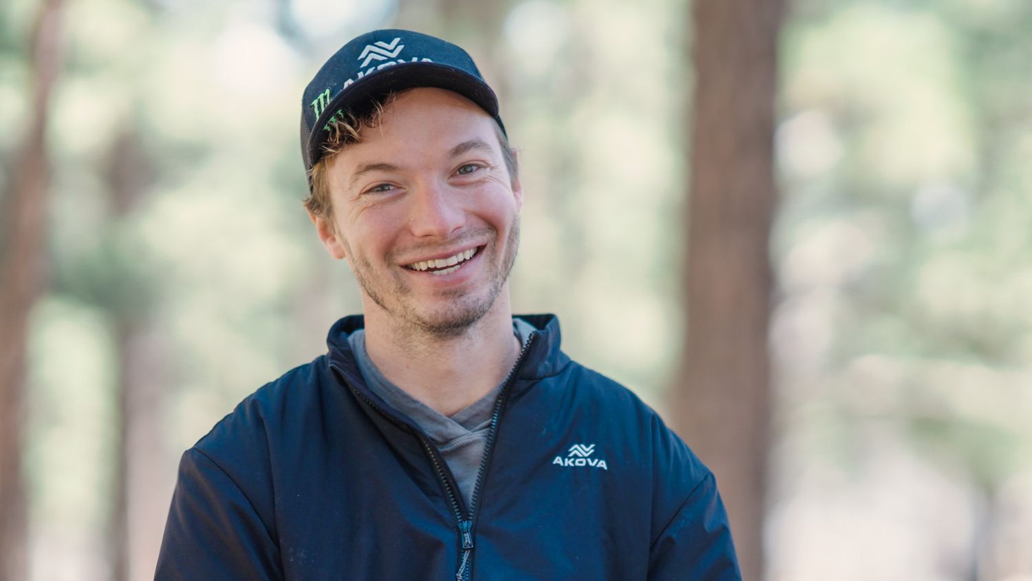 Person wearing navy cap and fleece jacket smiles outdoors in forest setting.