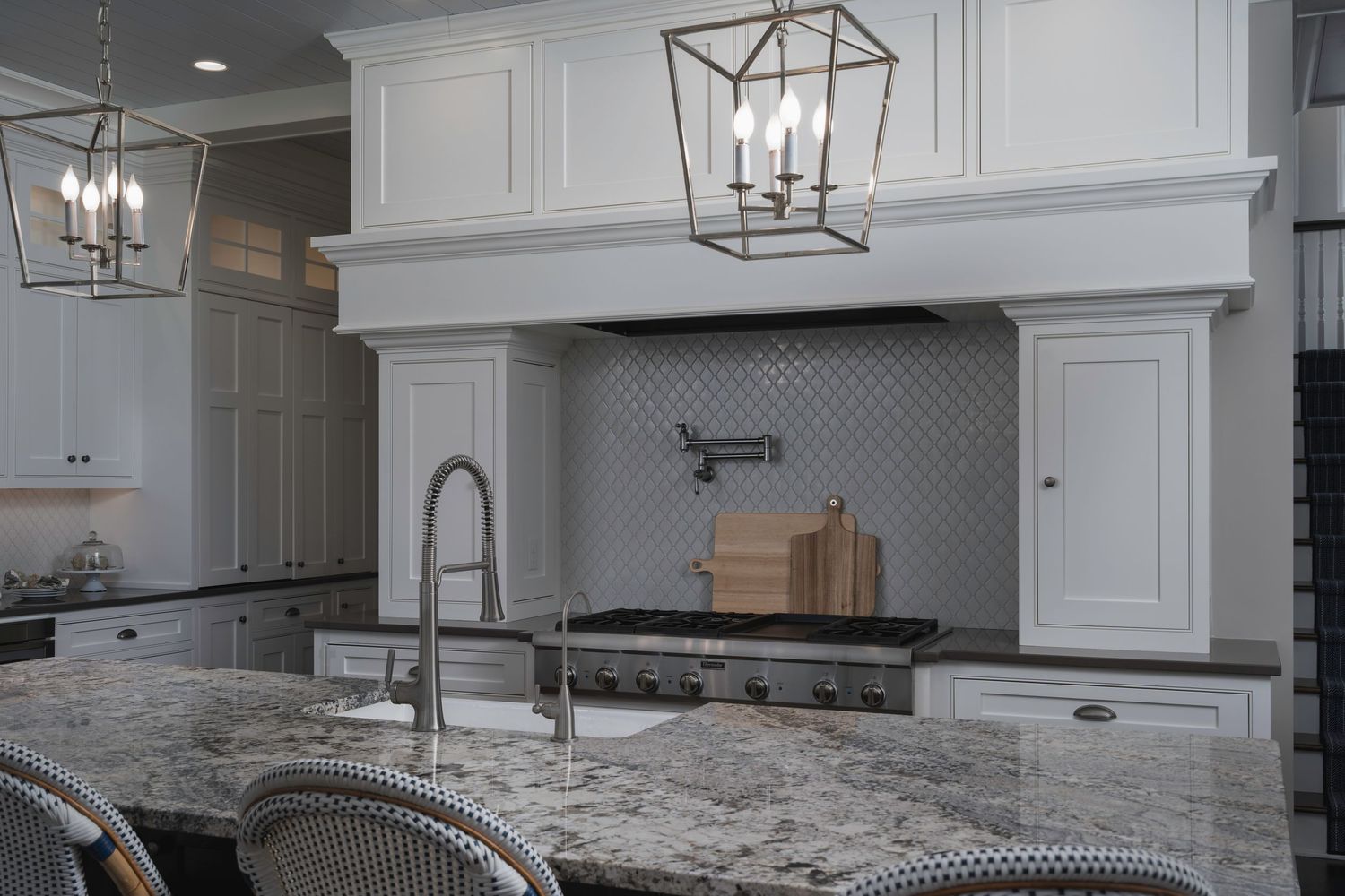 Elegant white kitchen with granite island, lantern pendant lights, stainless range, and decorative tile backsplash.