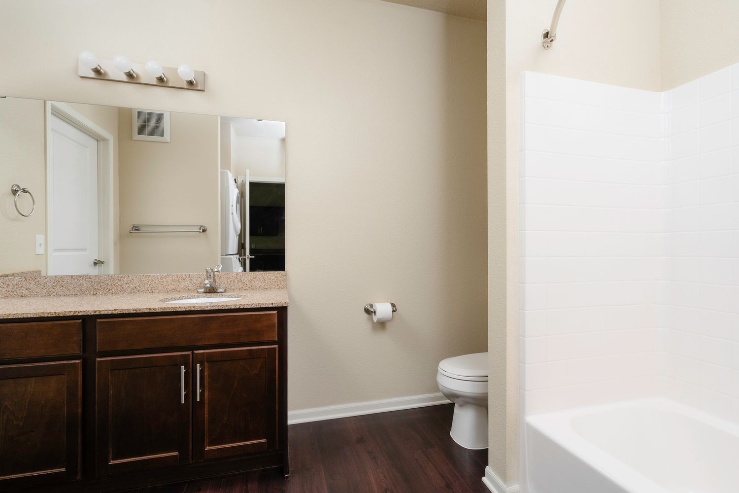 Modern bathroom with dark wood vanity, granite countertop, large mirror, white toilet, and soaking tub on hardwood floors.