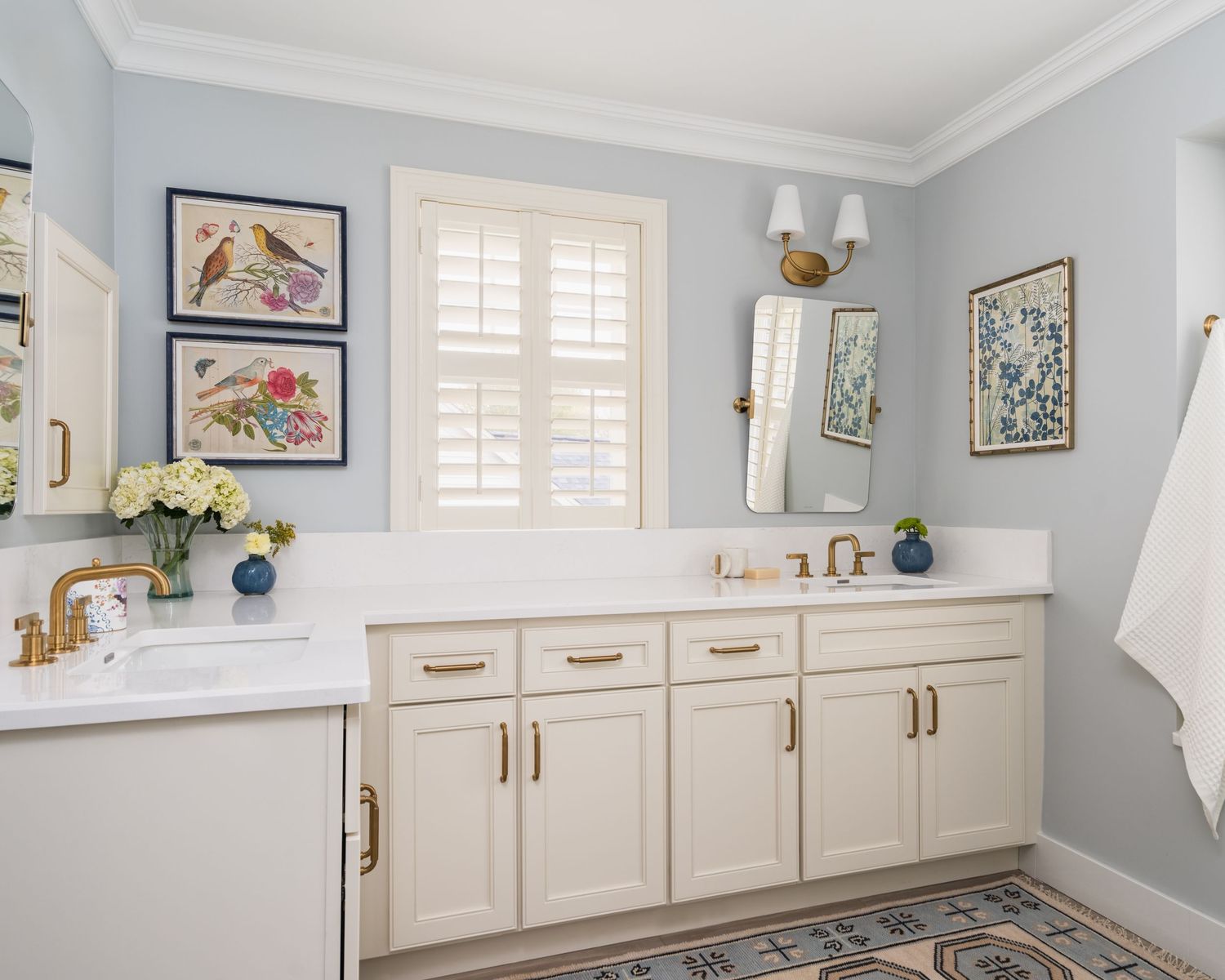 Elegant bathroom with white cabinetry, gold hardware, blue walls, botanical artwork, and a double vanity with marble countertops.