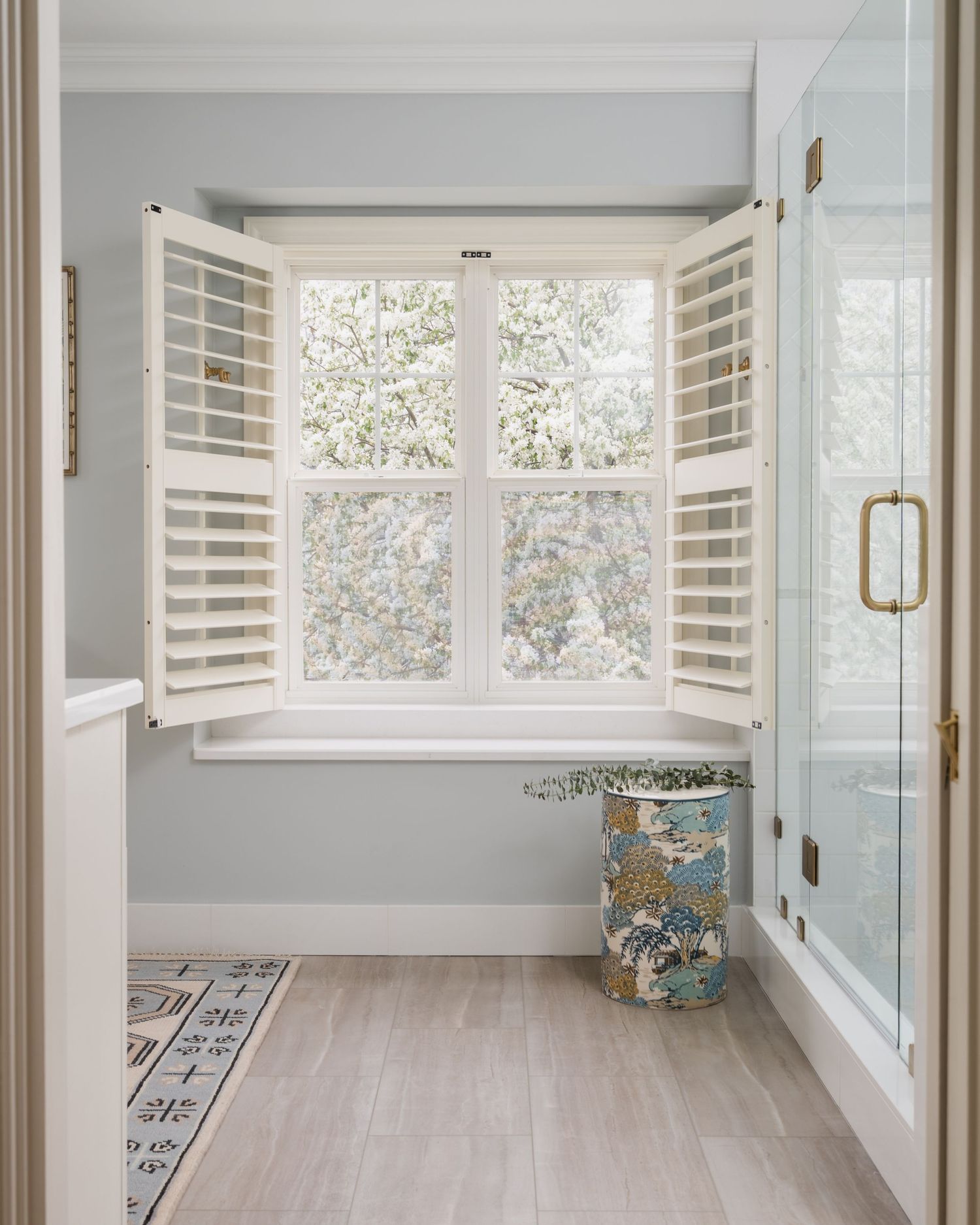Bright bathroom with open white plantation shutters, light gray walls, wood-look floors, floral waste bin, and glass shower door.