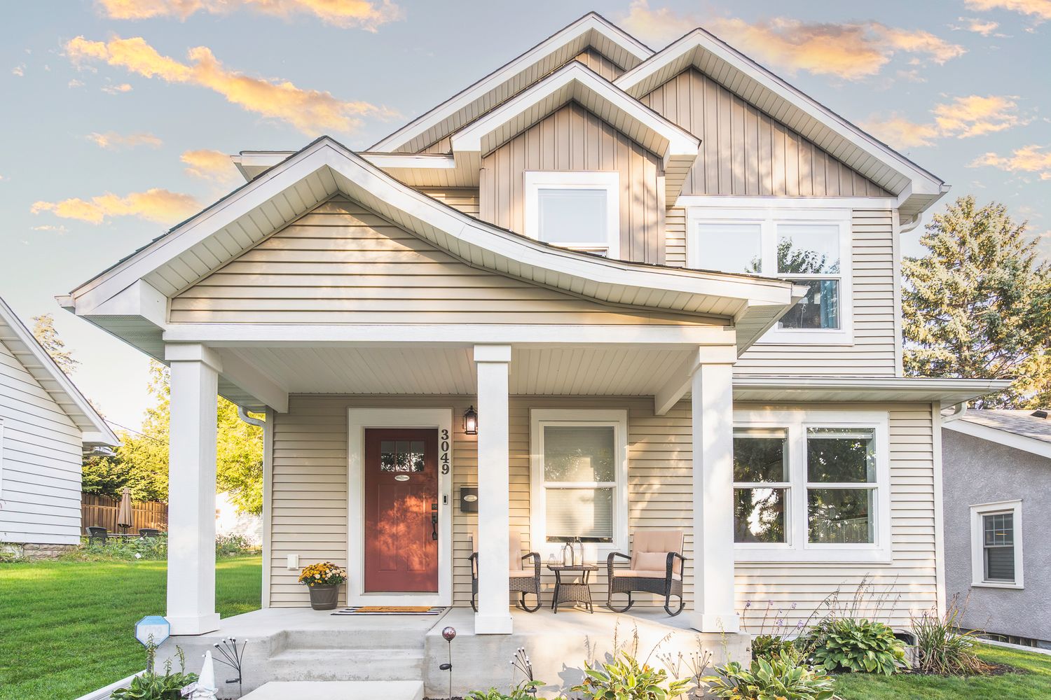 Charming two-story craftsman home with covered front porch, beige siding, red door, and lush landscaping at sunset.