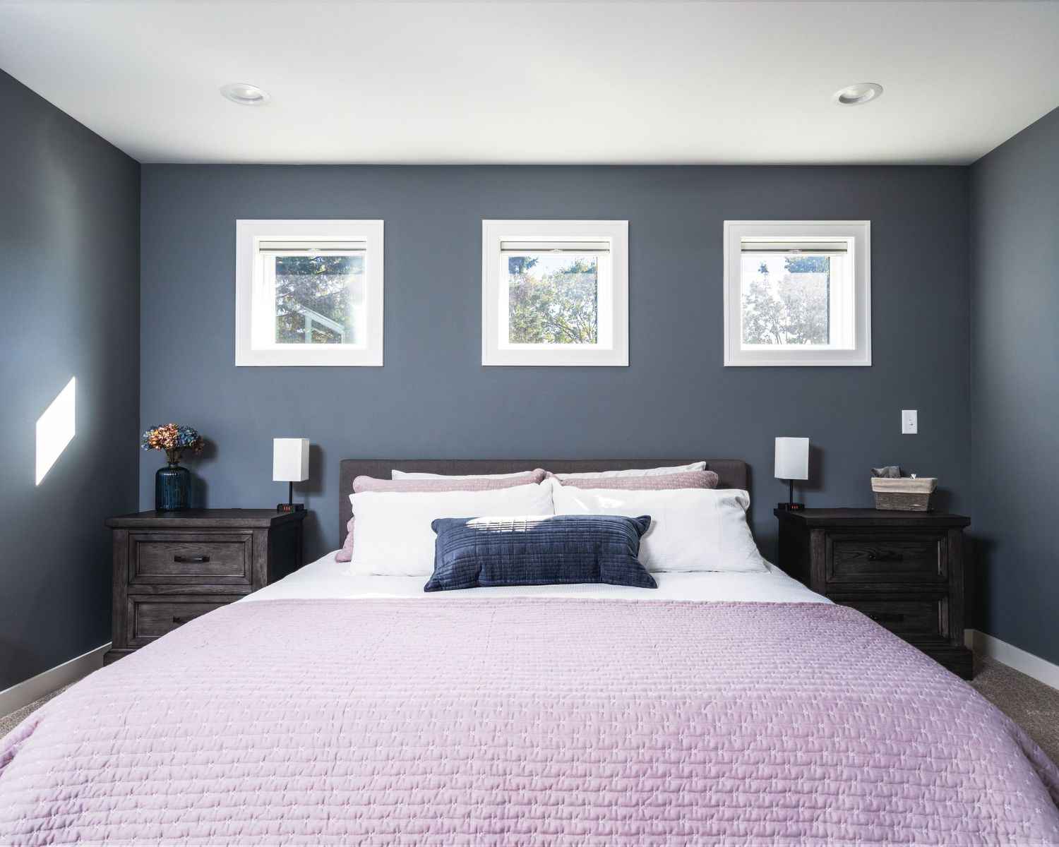 A modern bedroom with dark blue walls features white trim and a light pink bedspread with natural light from windows.