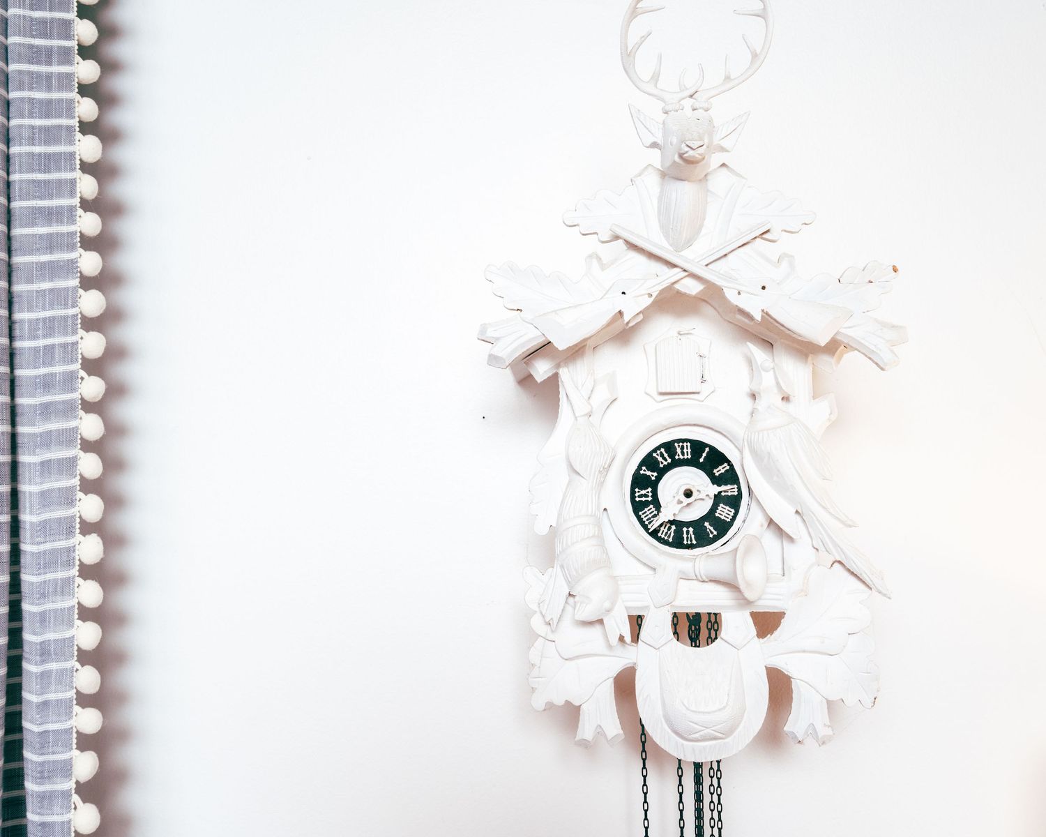 A white decorative cuckoo clock with deer antlers mounted on a light colored wall next to a spiral notebook.