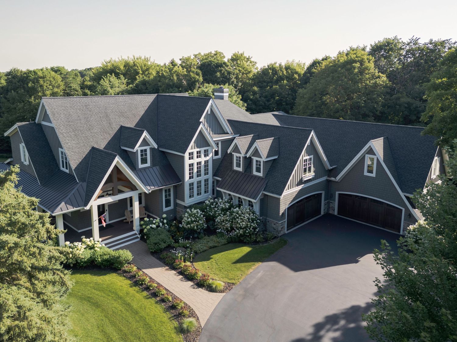 Aerial view of a large luxury gray shingle home with dark roof, attached garage, and lush green tree surroundings.