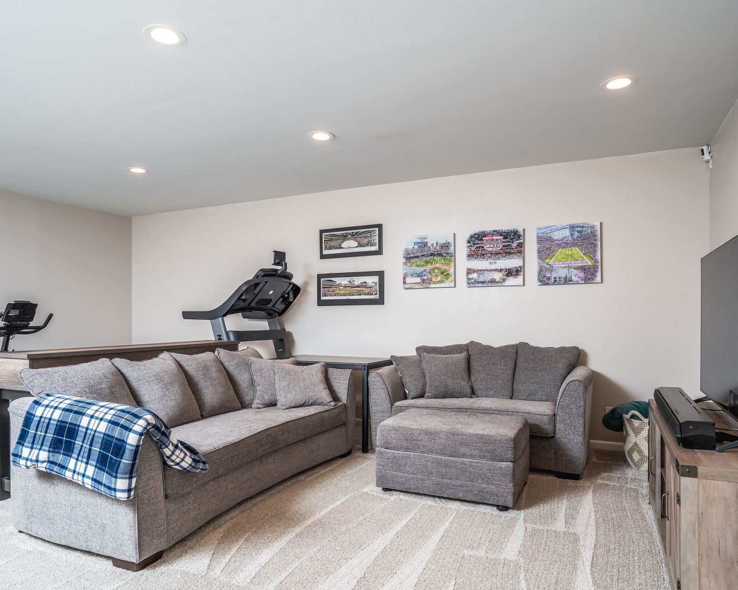 Modern basement living room with gray sectional sofa, exercise equipment and framed artwork on white walls.