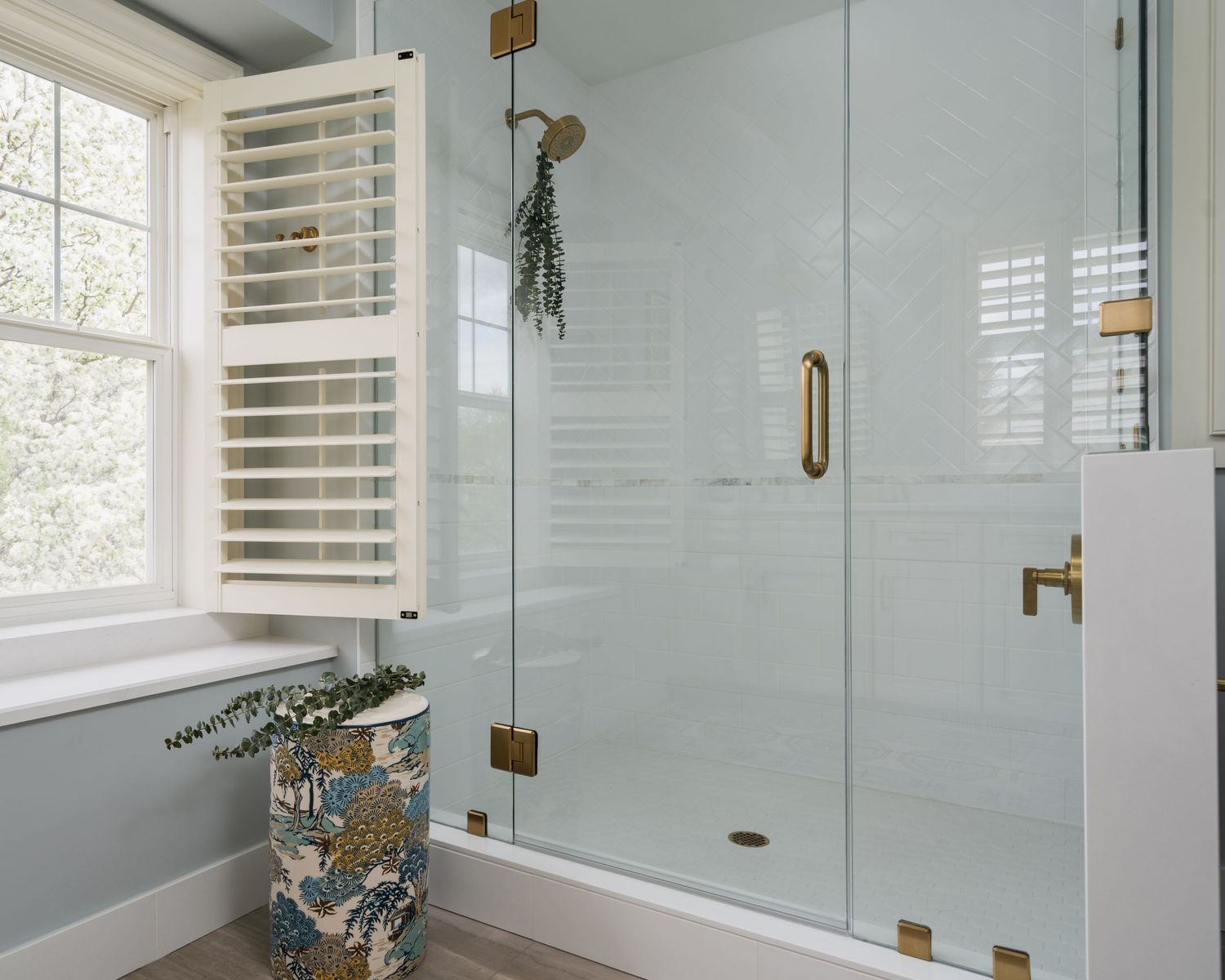 Modern bathroom with frameless glass shower, brass hardware, plantation shutters, and decorative laundry basket near window.