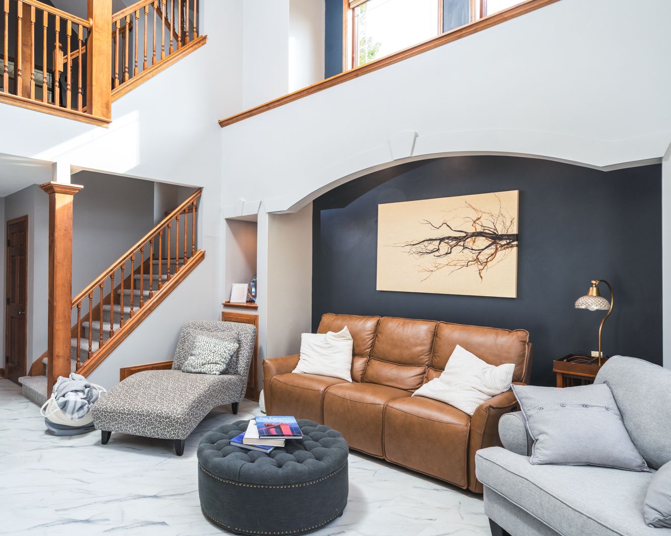 Modern living room with tan leather sofa, gray chairs, dark accent wall, and wooden staircase in open concept space.
