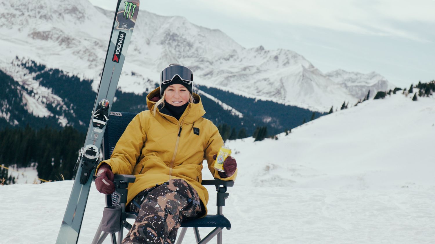Person in yellow jacket sits in camping chair with skis on snowy mountain slope.