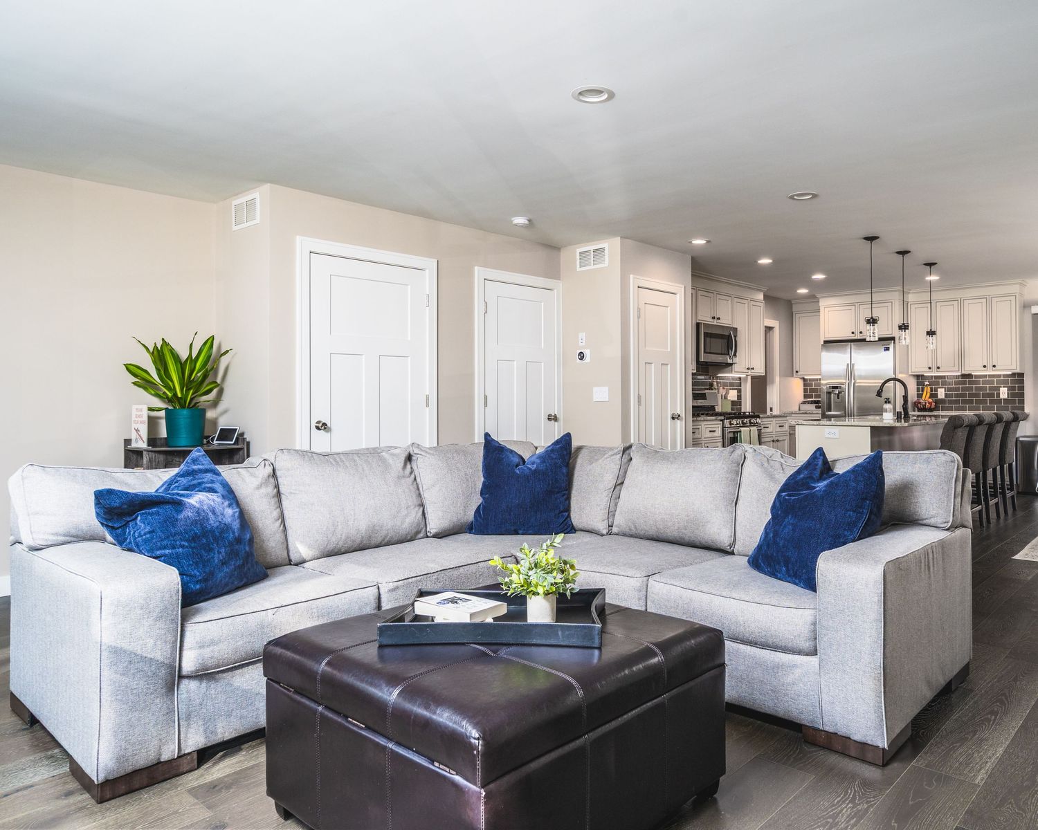 Modern living room features white sofas with navy blue accent pillows and a dark leather ottoman in an open concept space.