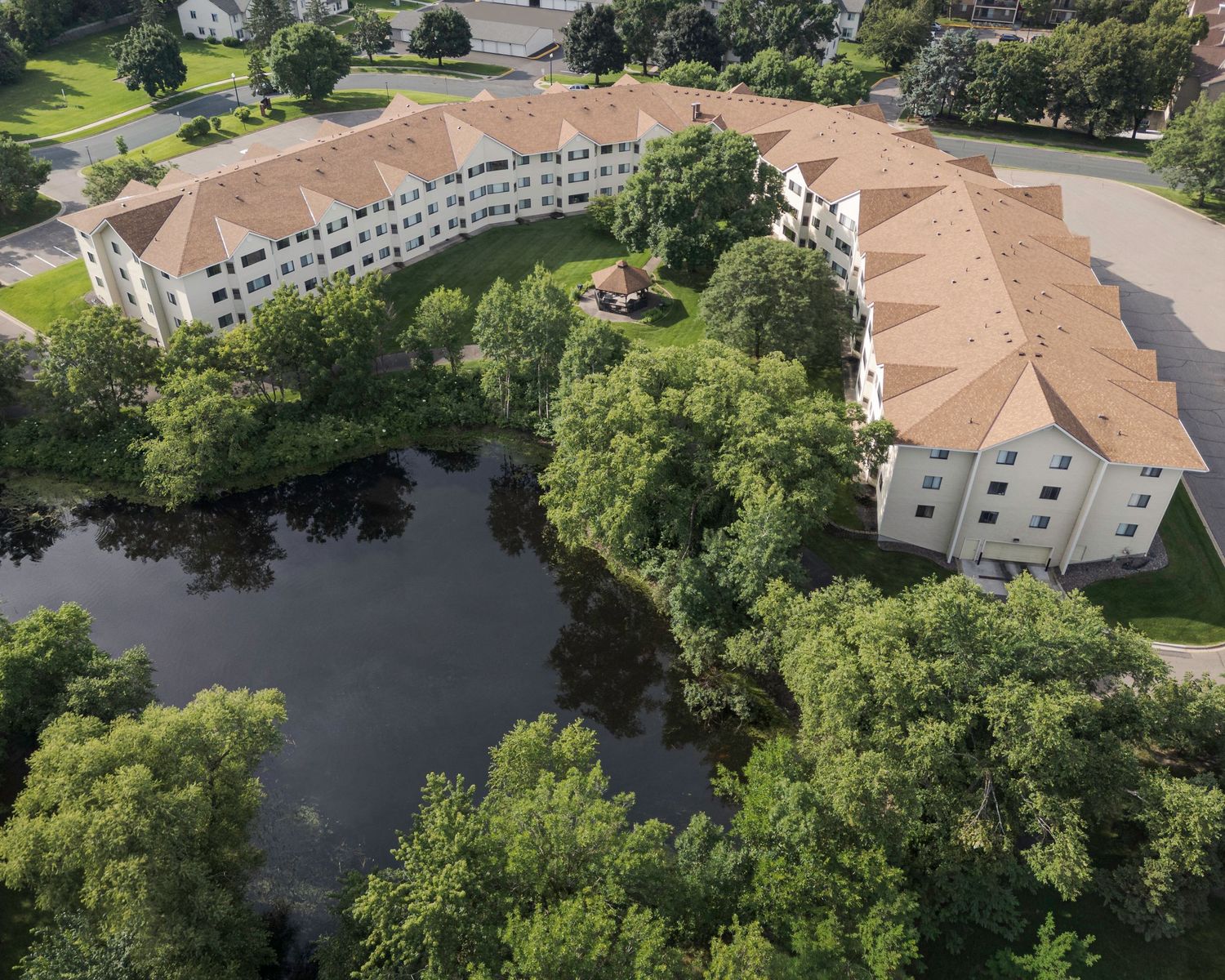 Aerial view of a curved apartment complex with orange roofs surrounded by lush trees and a serene pond.