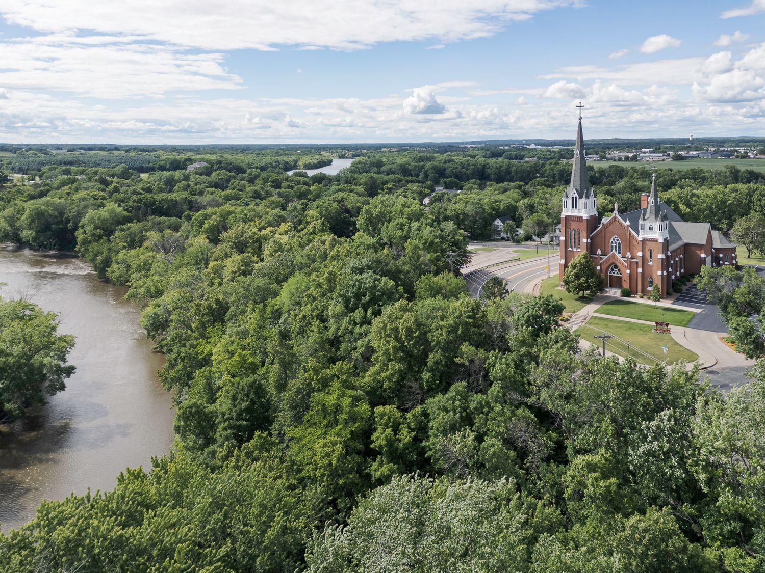 Aerial view of a red brick church with a tall steeple surrounded by lush green trees and a river in a small town.