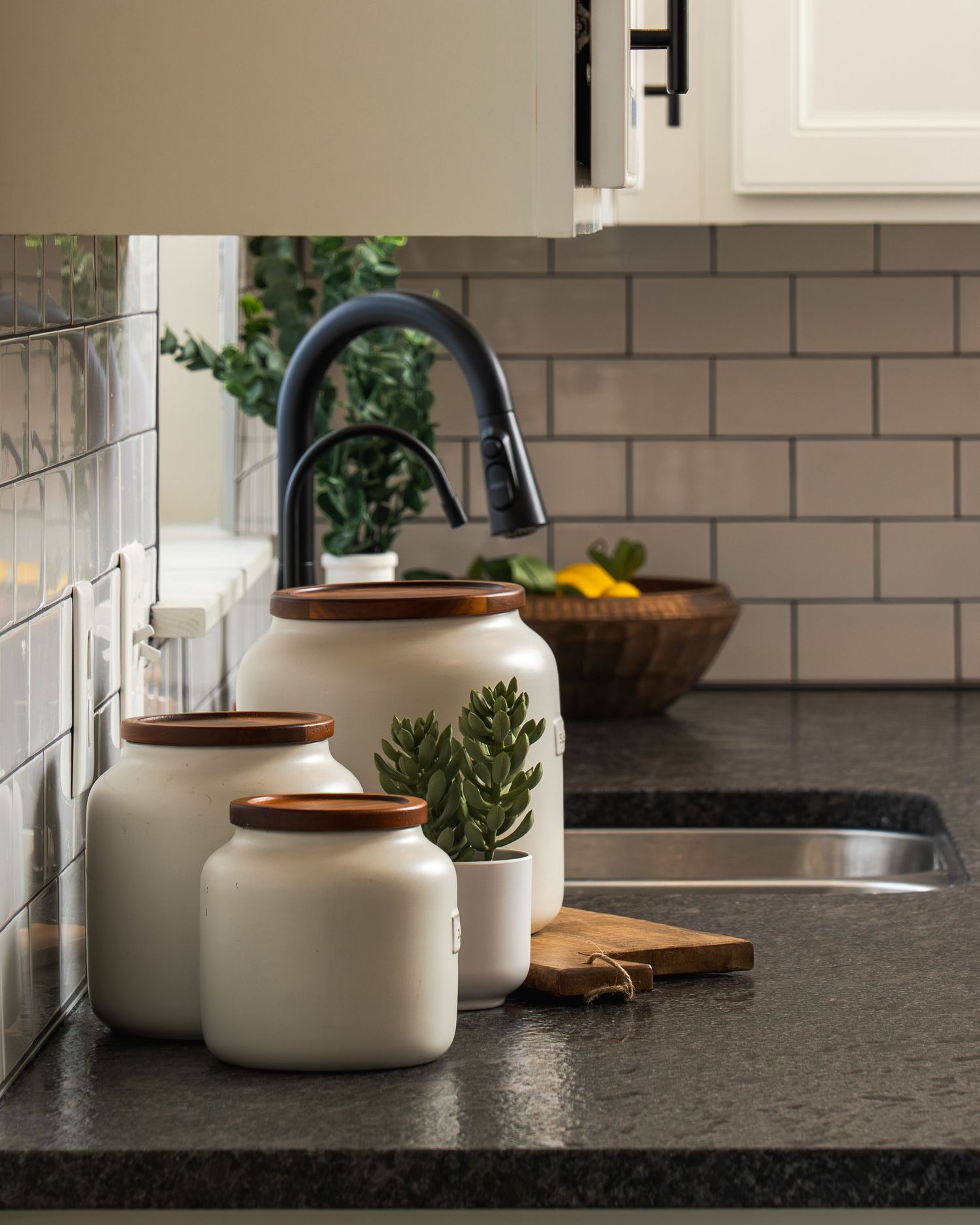 Modern kitchen counter with black faucet, ceramic canisters, and subway tile backsplash against dark granite countertop.