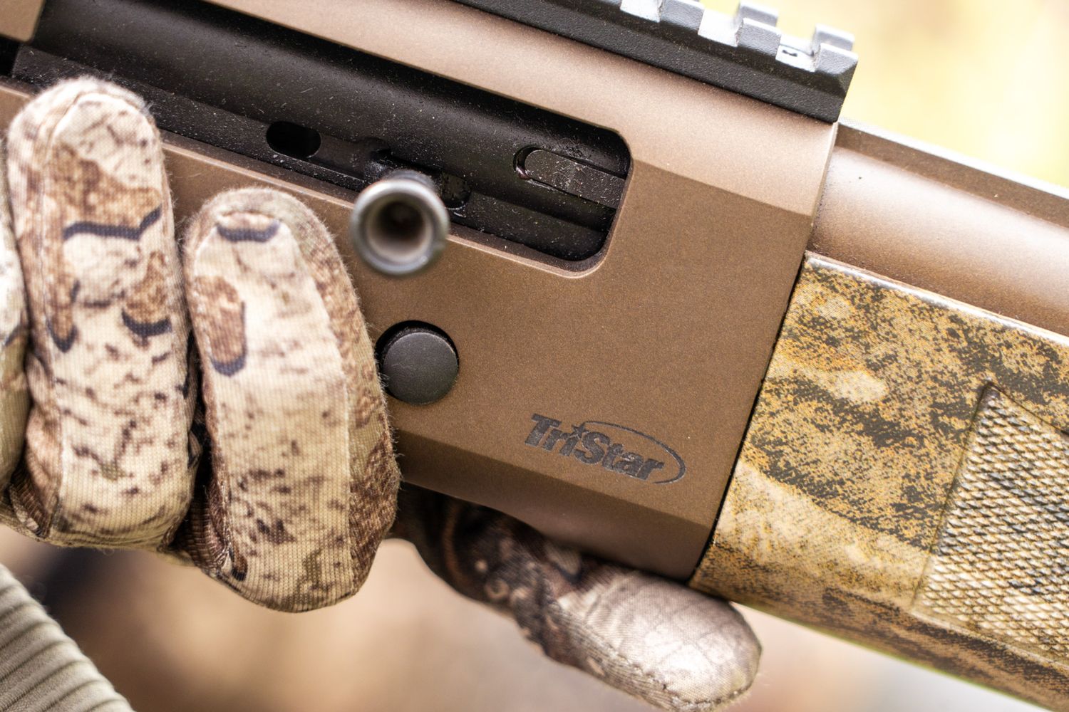 Close-up of camouflage gloved hands holding tan tactical shotgun.